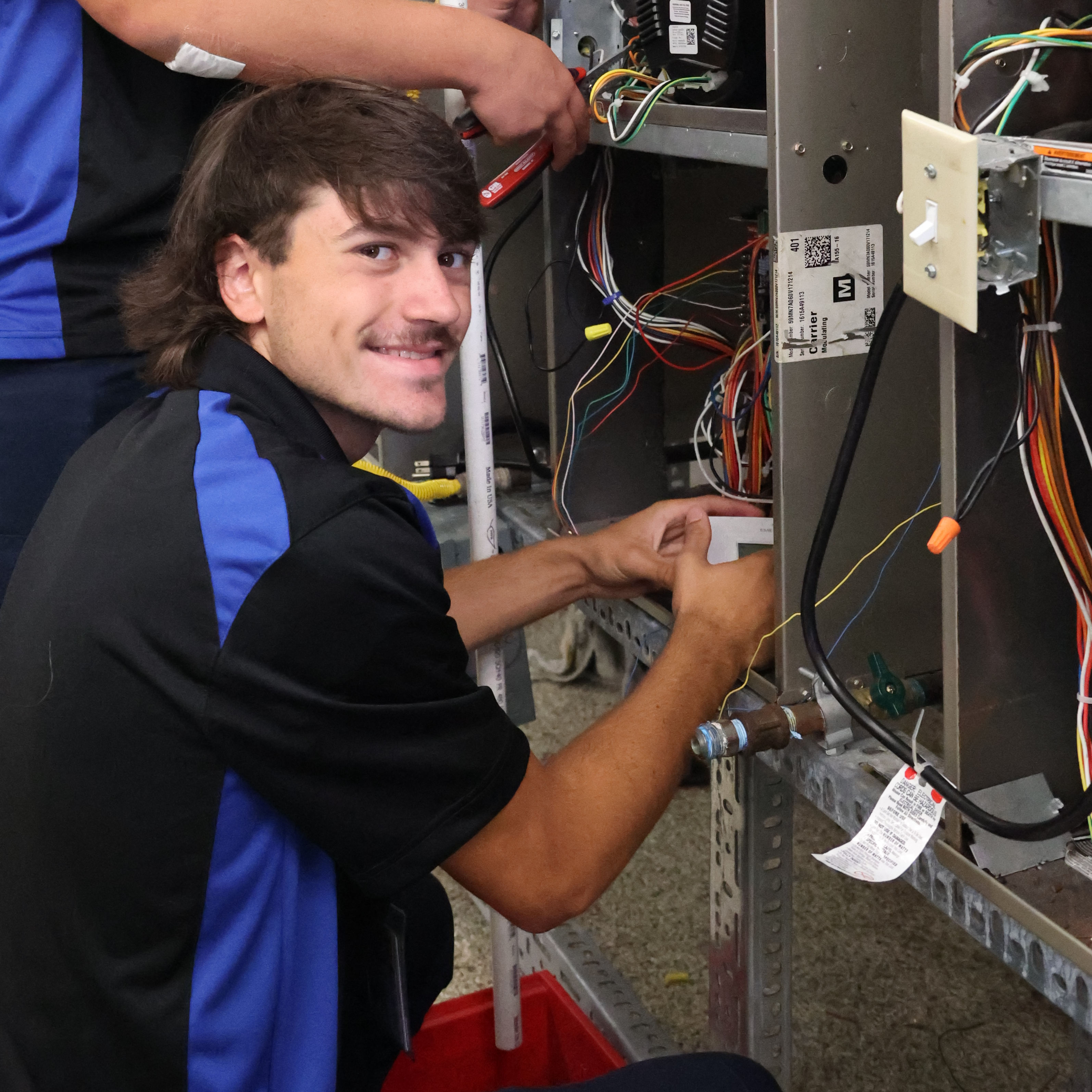 Two White, male students have the back of air conditioning unit open, but pause to smile for a photo.