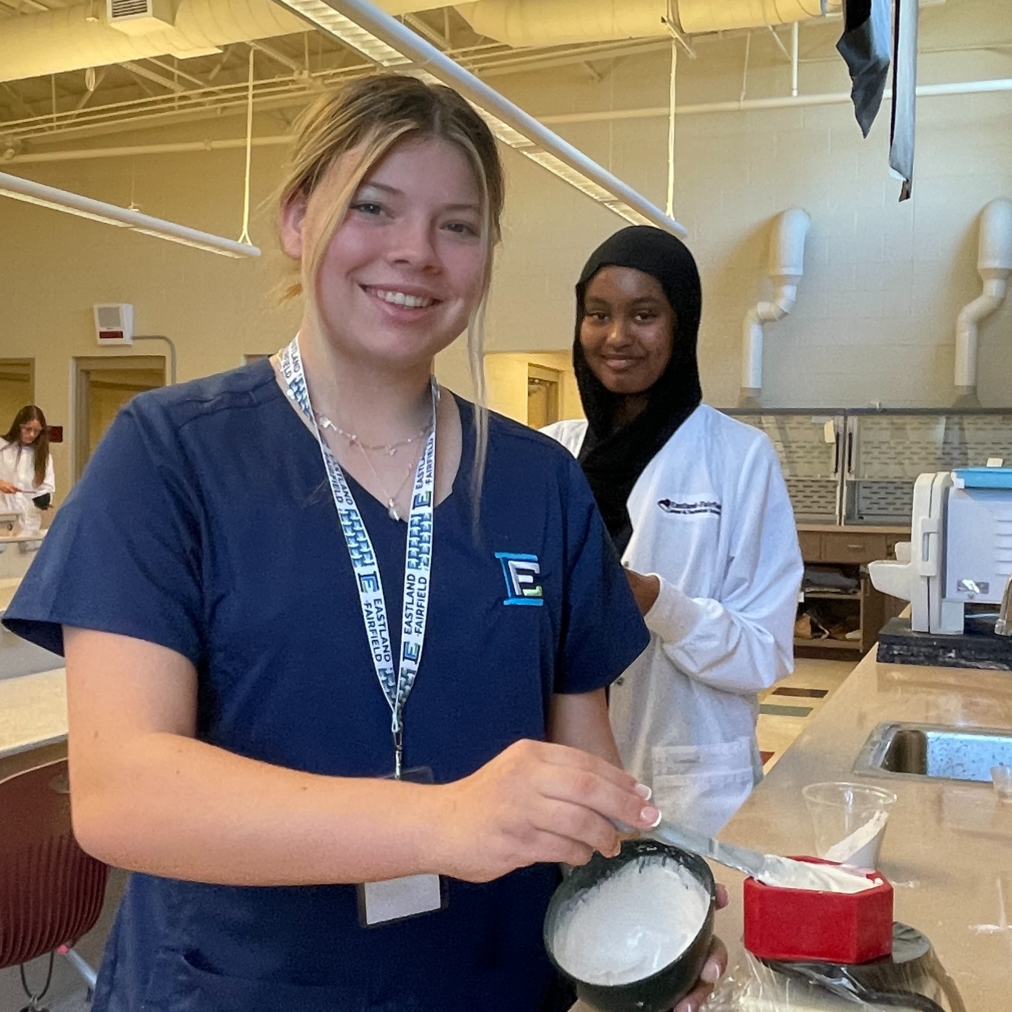 Two female students, one White and one Black wearing a hijab, smile while they finish mixing their putty impression kits.