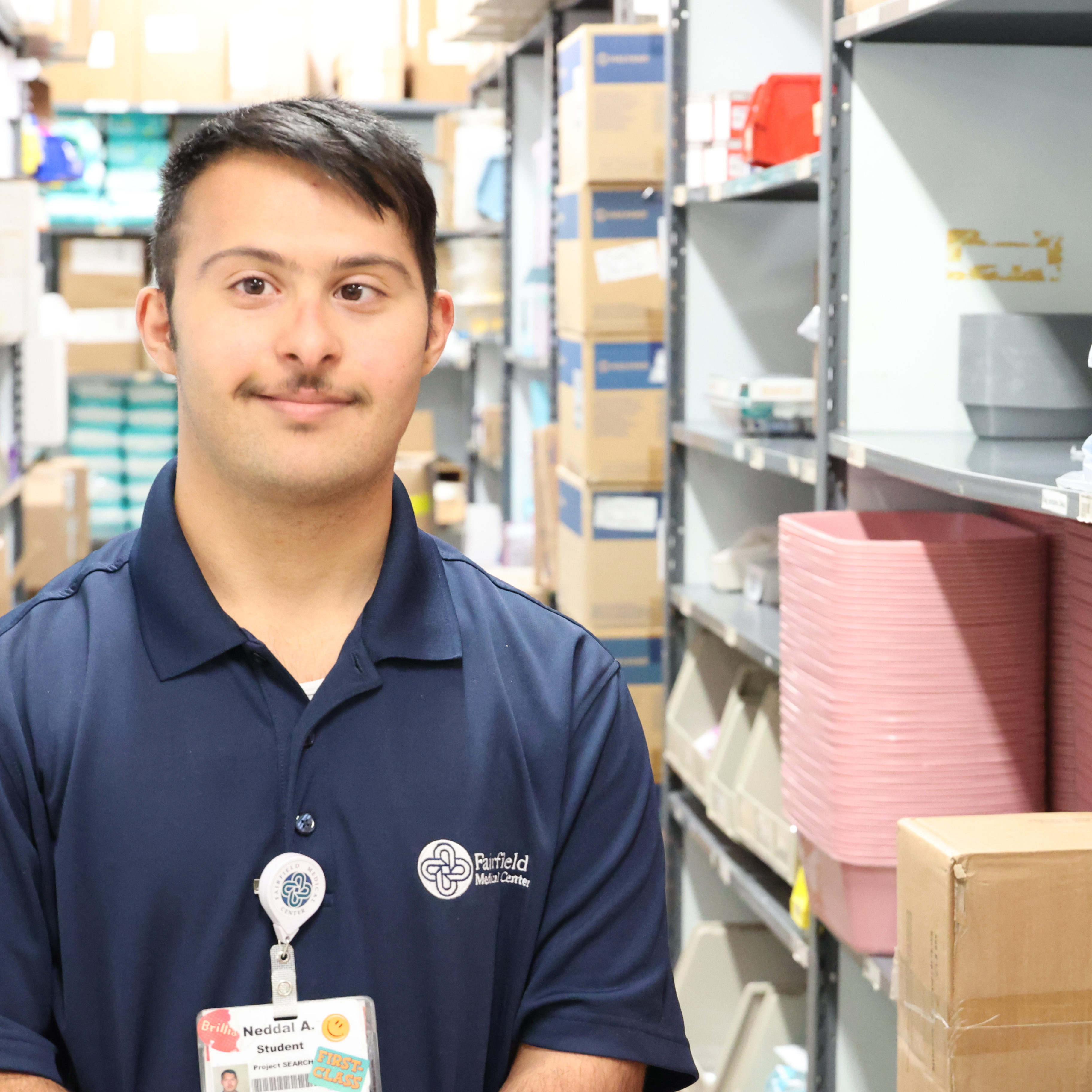 A Hispanic, male student wears a Fairfield Medical polo shirt in the aisle of a stockroom.