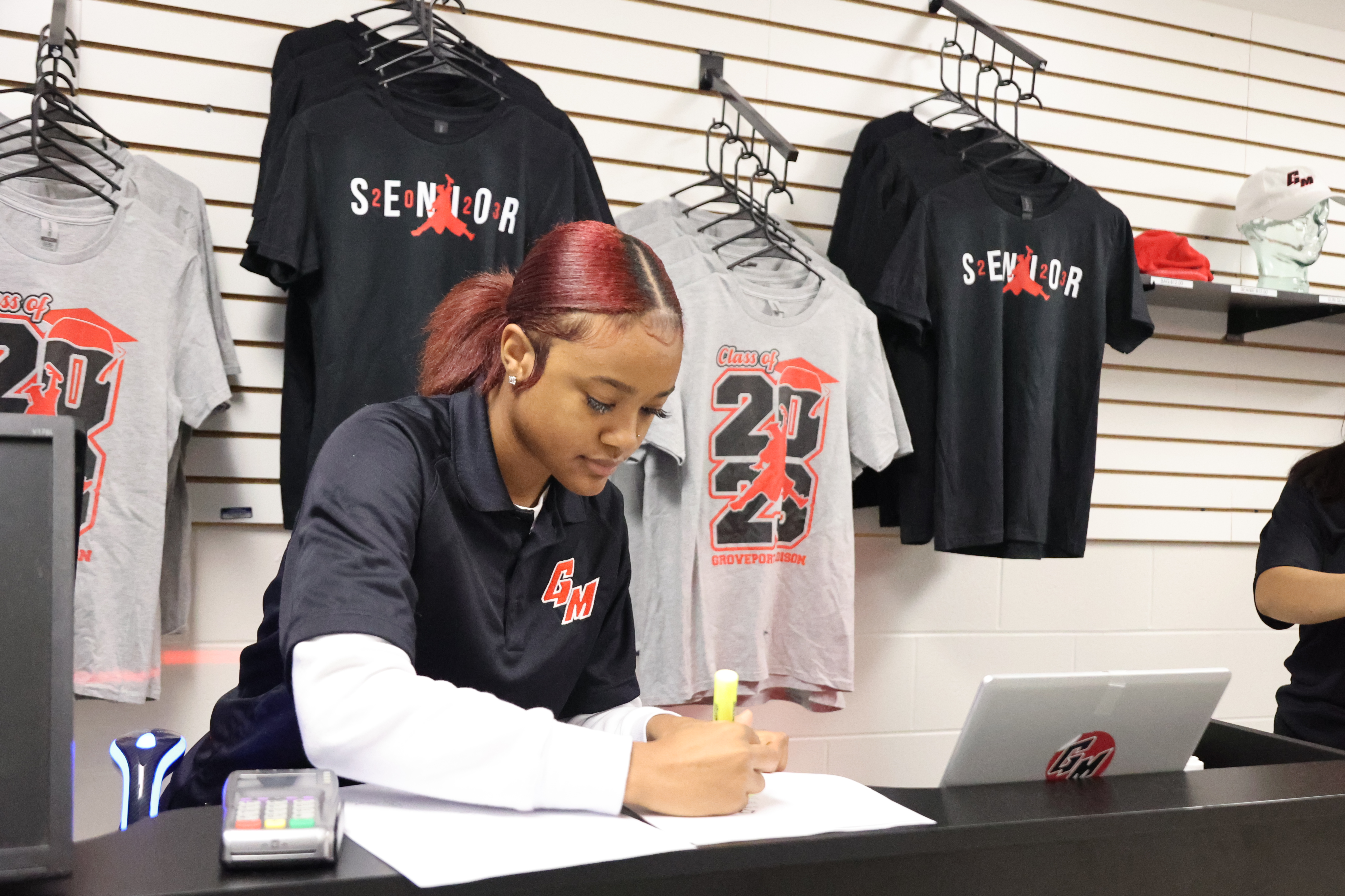 A Black, female is doing paperwork near a cash register and in front of t-shirts hanging on the wall.