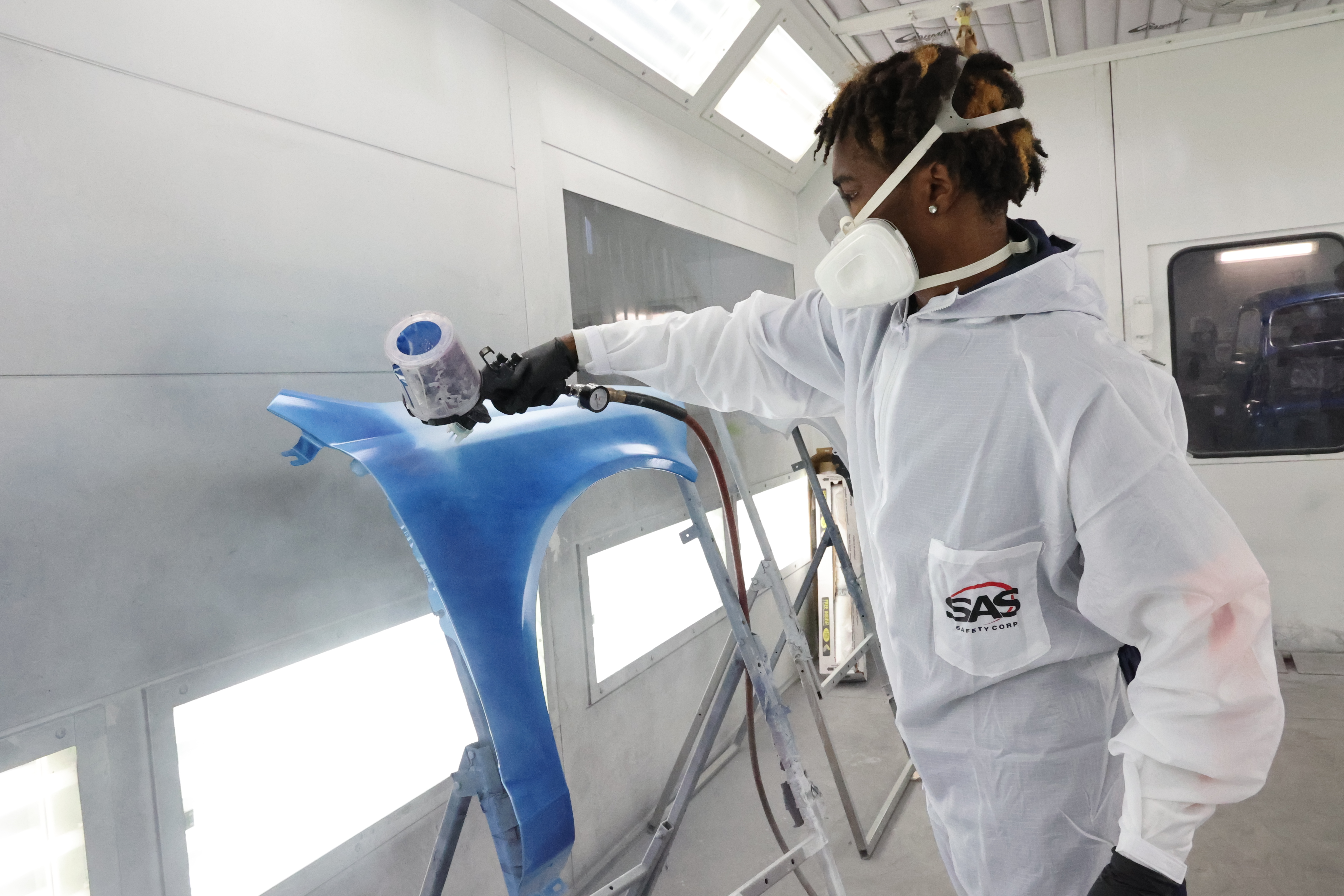 A Black, male student is in a white paint suit and applying blue paint to a fender.