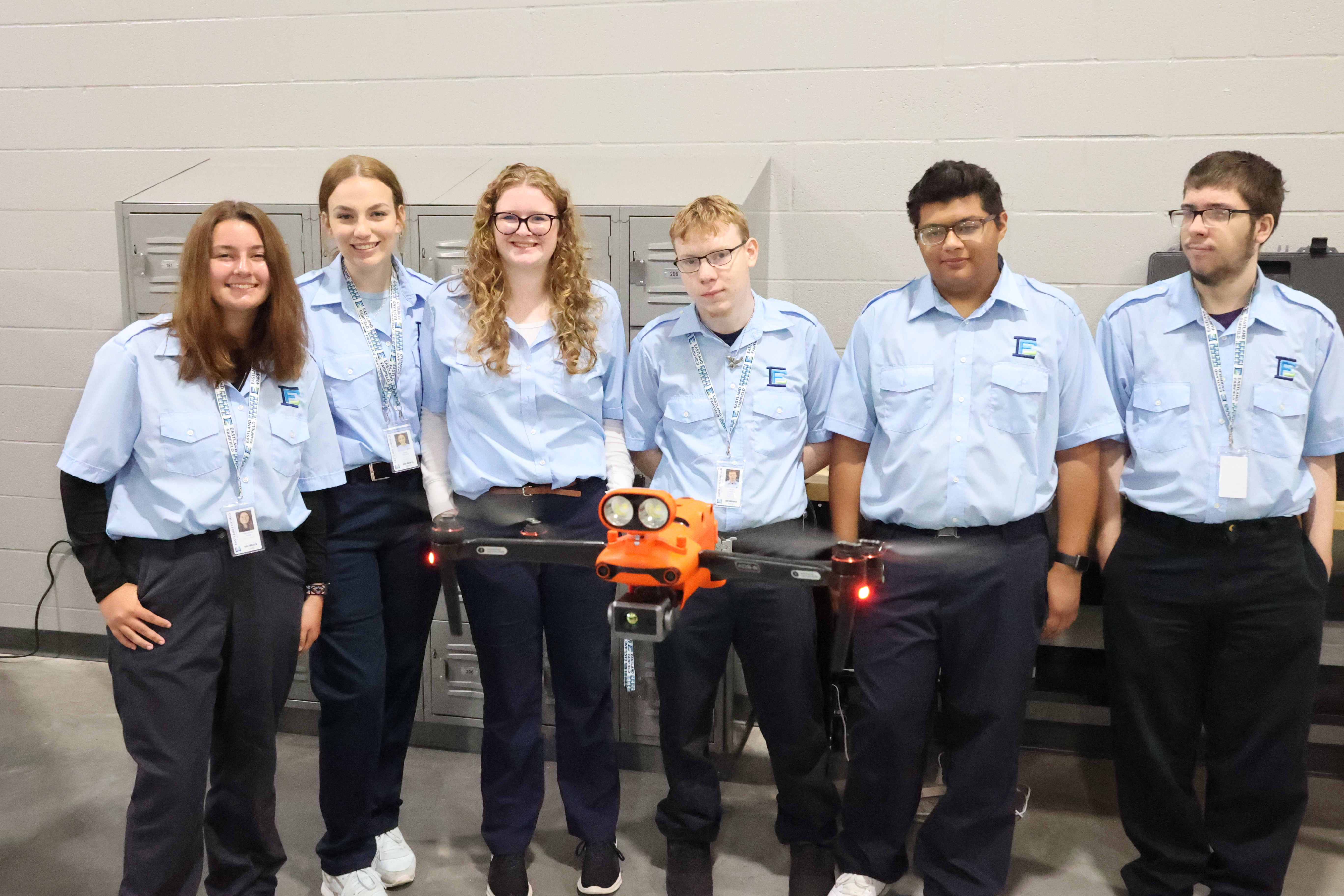 A group of five students of mixed backgrounds lines up behind a drone that is flying in front of them.