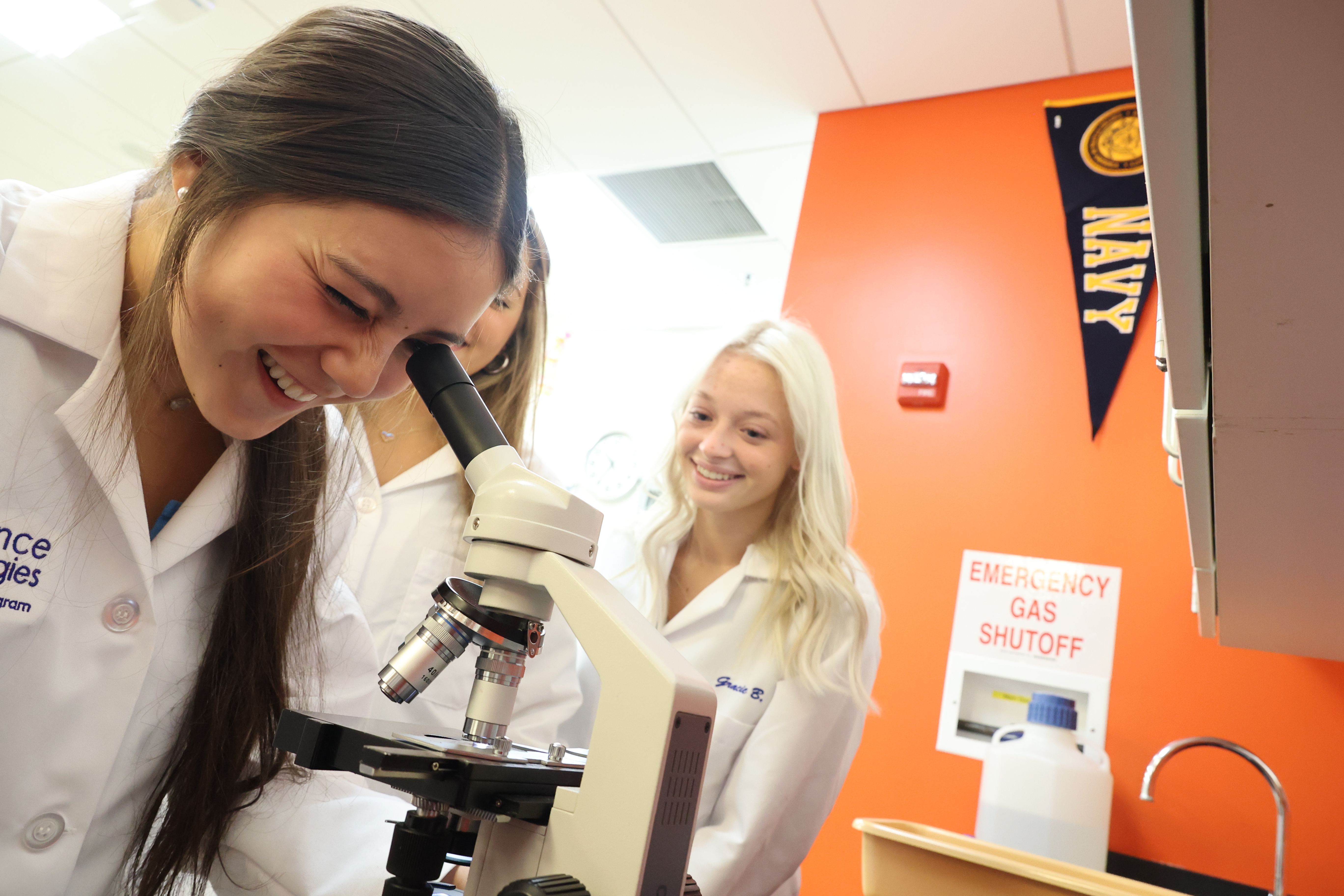 A group of female students gathers around a microscope, one peering down the eye piece.