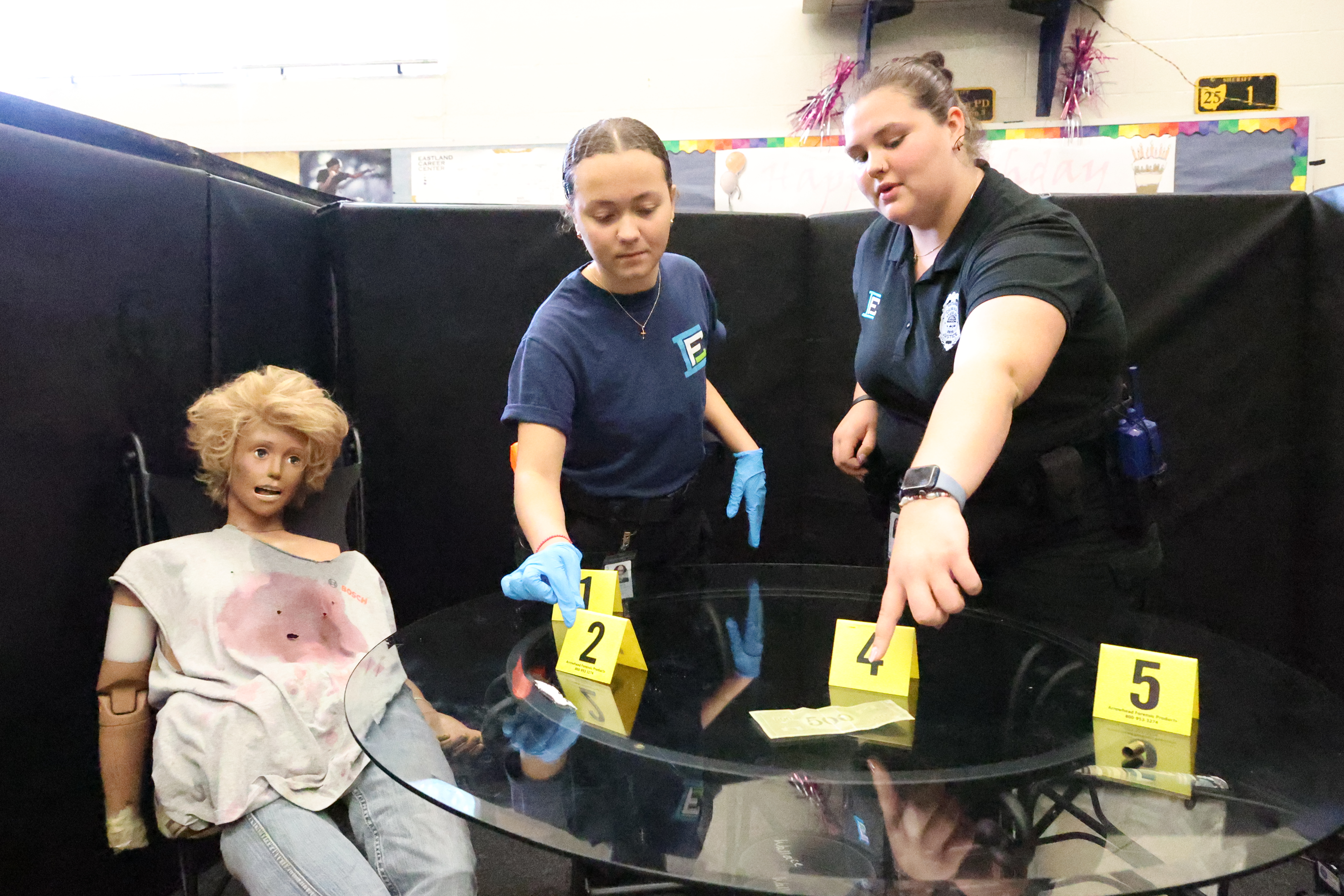 Two female students are examining a crime scene with a blood-soaked mannequin posed as a victim.