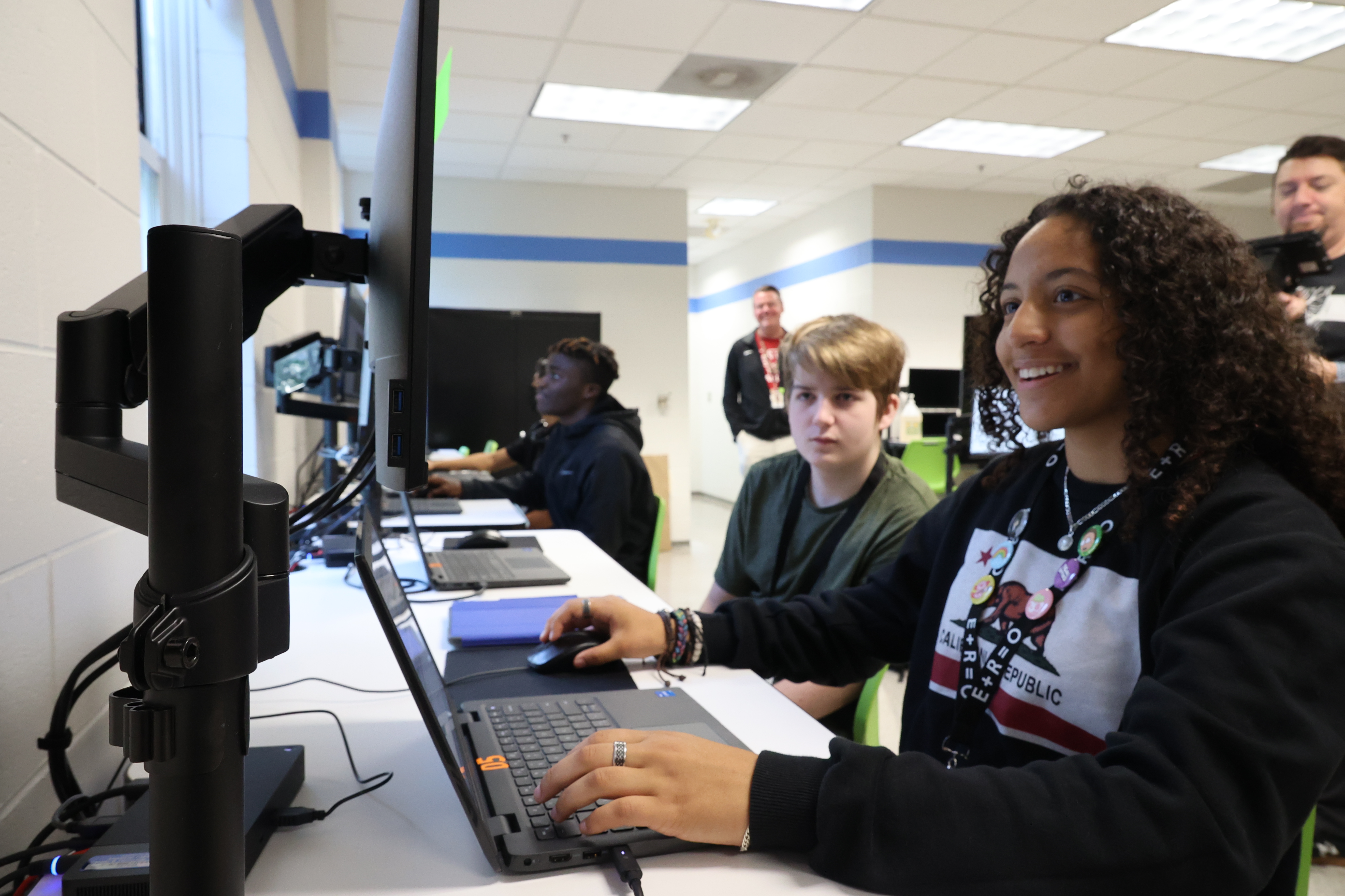 A Black, female student smiles as she types code on a desktop computer with a classmate looking over her shoulder.