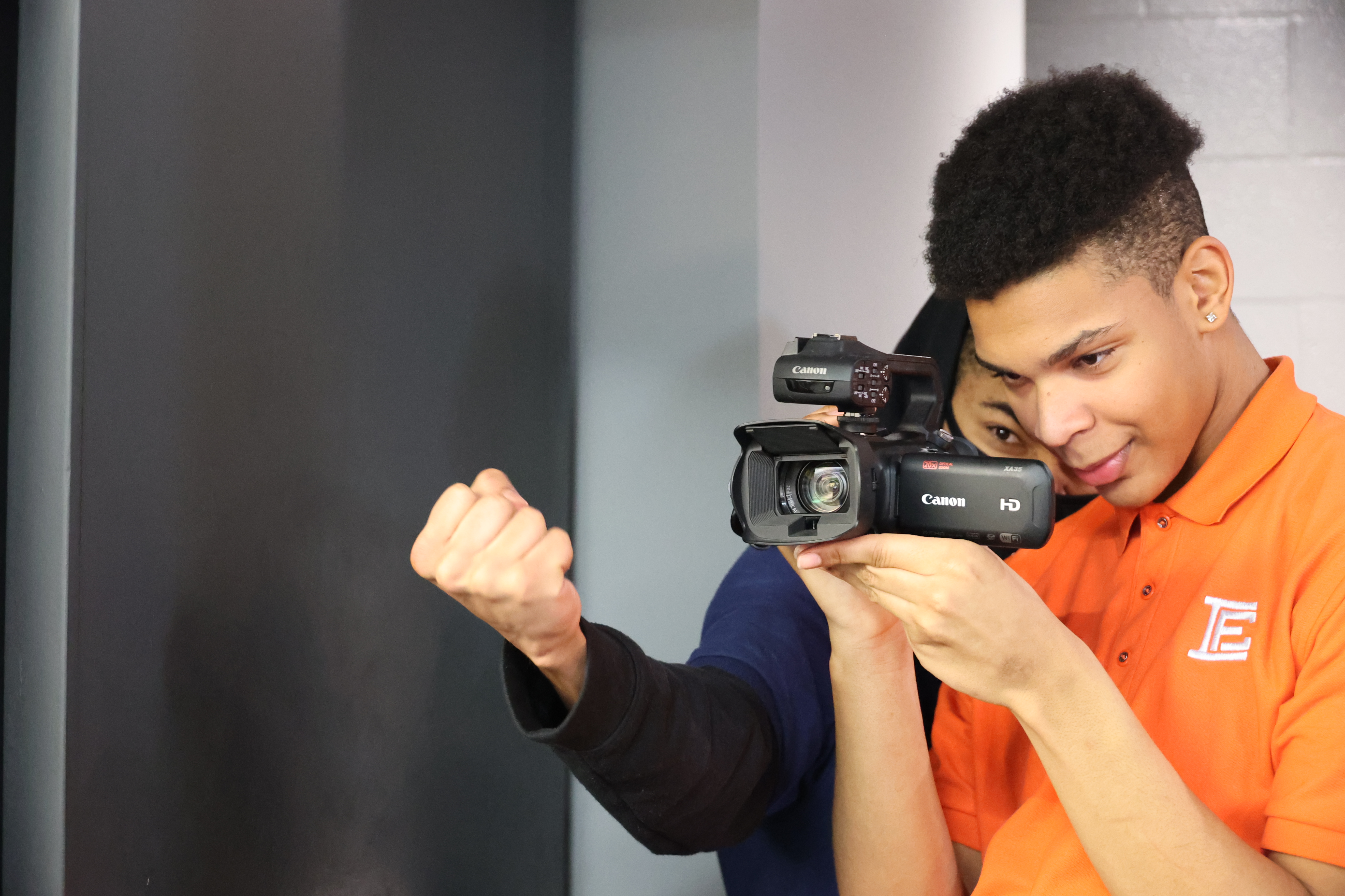 A student is holding a Canon camera to film another student raising a fist in front of the lens to capture the shot.