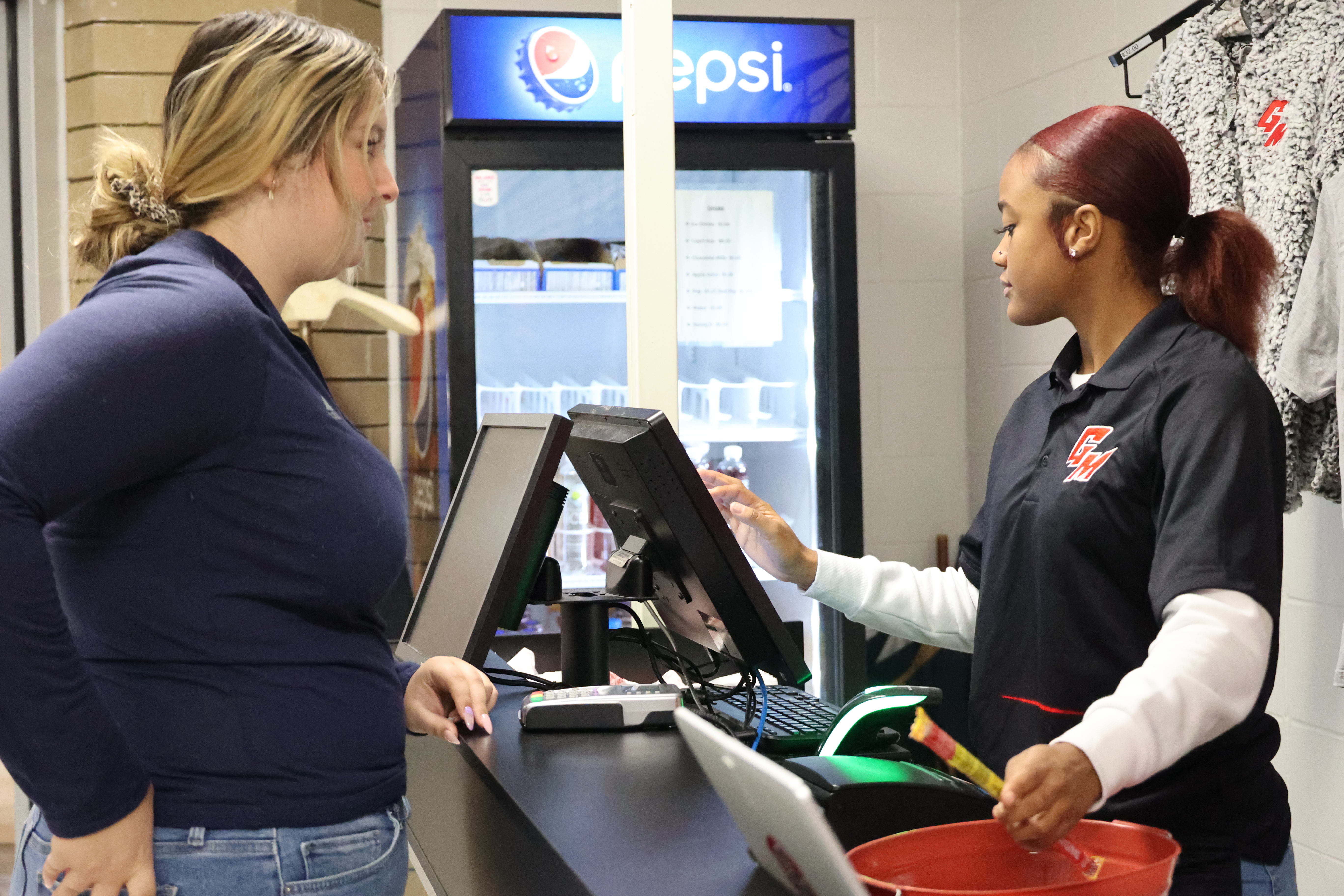 A Black, female student assists another White student at the cash register.