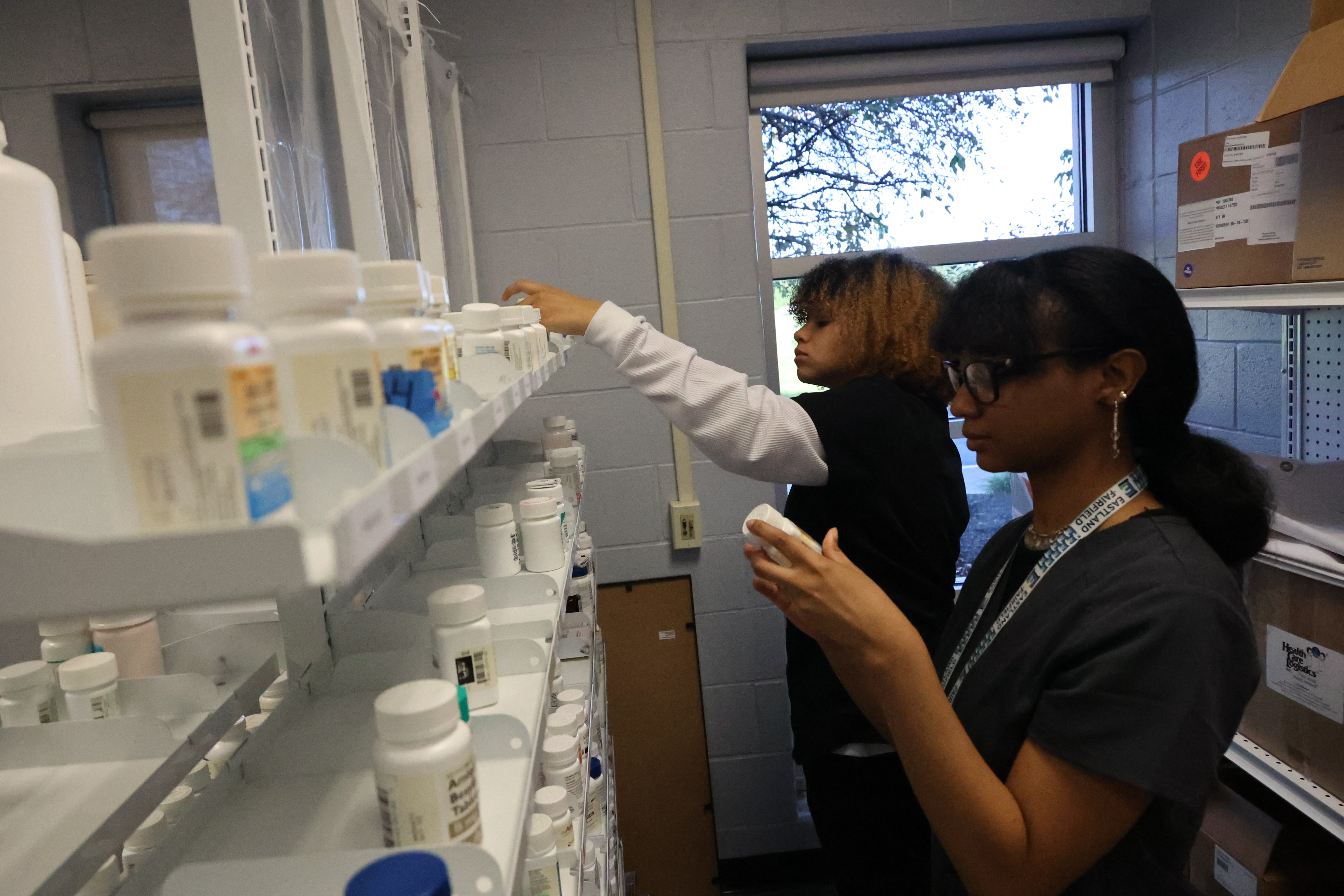 Two students are examining and sorting placebo medication on the shelves of the lab pharmacy