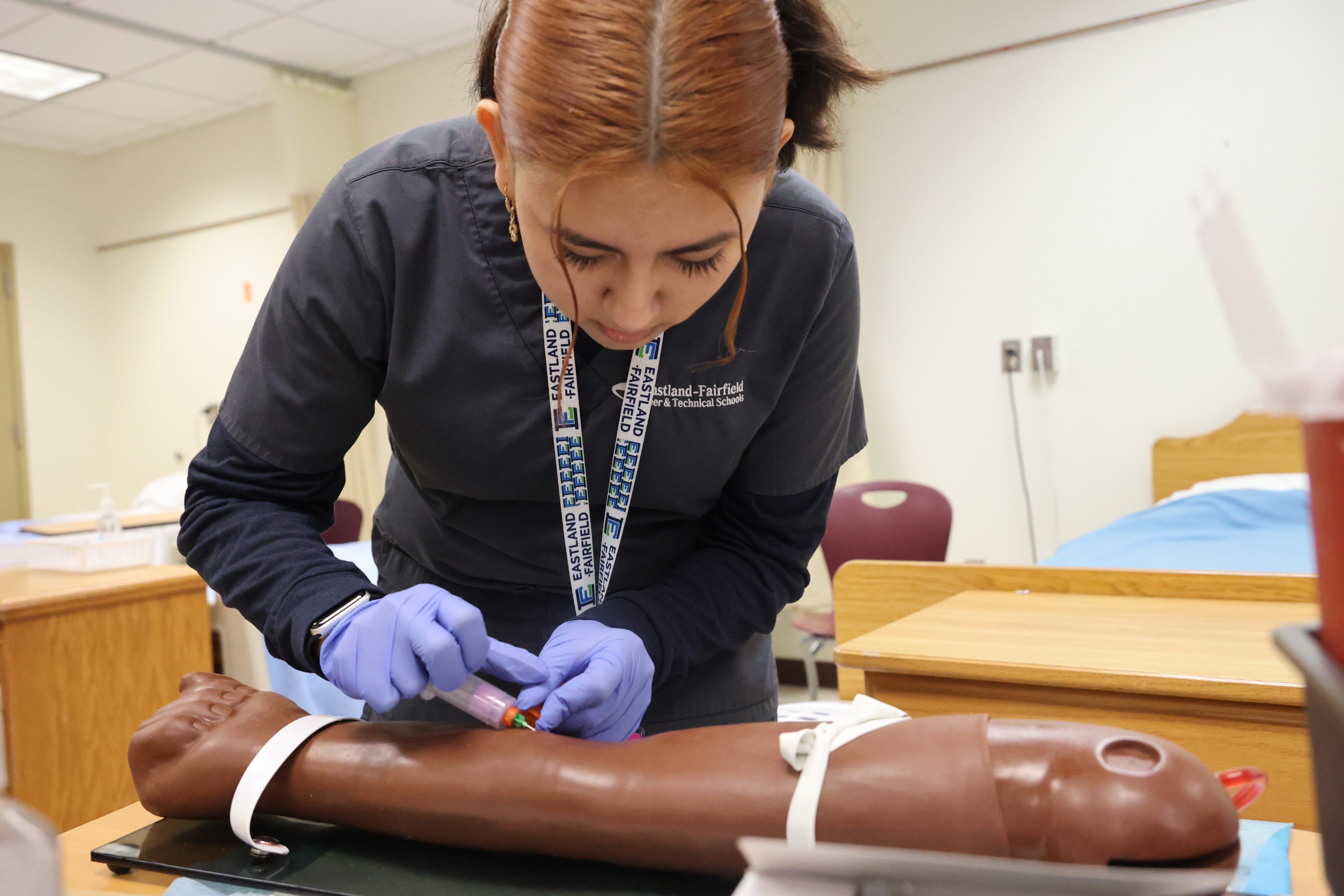 A female student is penetrating a blood drawing needle into a dark colored mannequin arm.