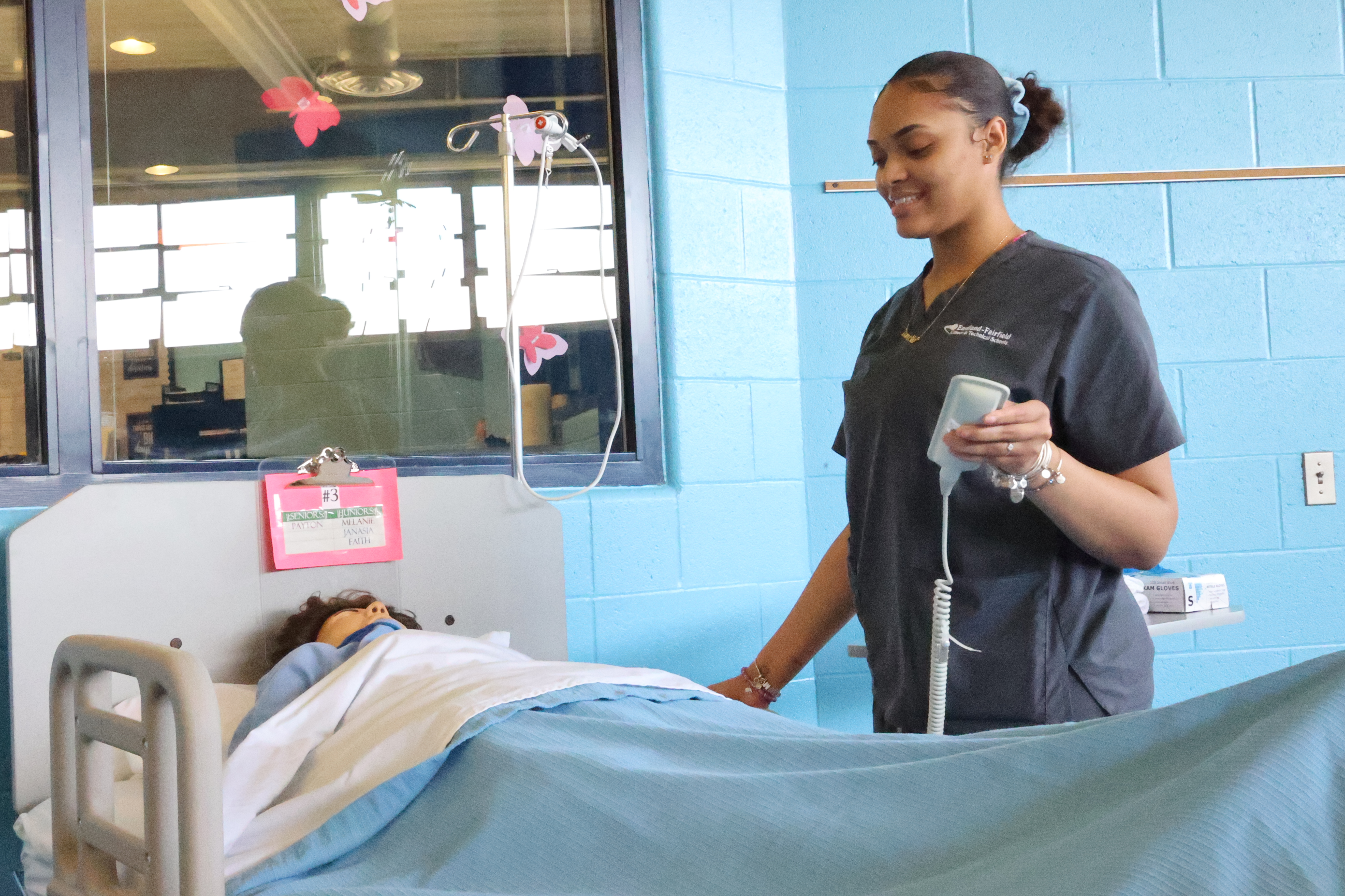 A Black, female student greets her patient while holding the bed remote control.