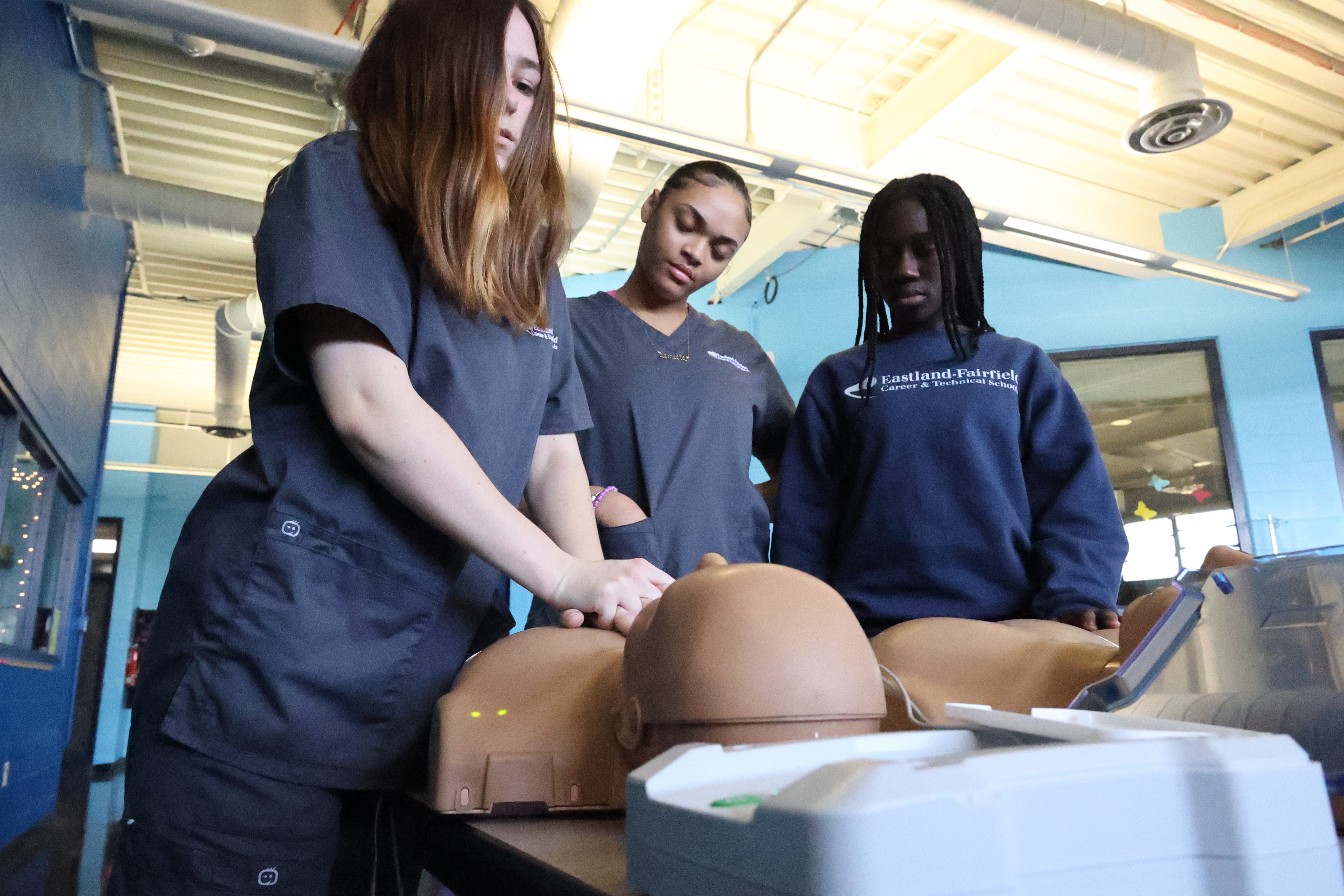 A group of students huddle around CPR training devices as one student applies compressions.