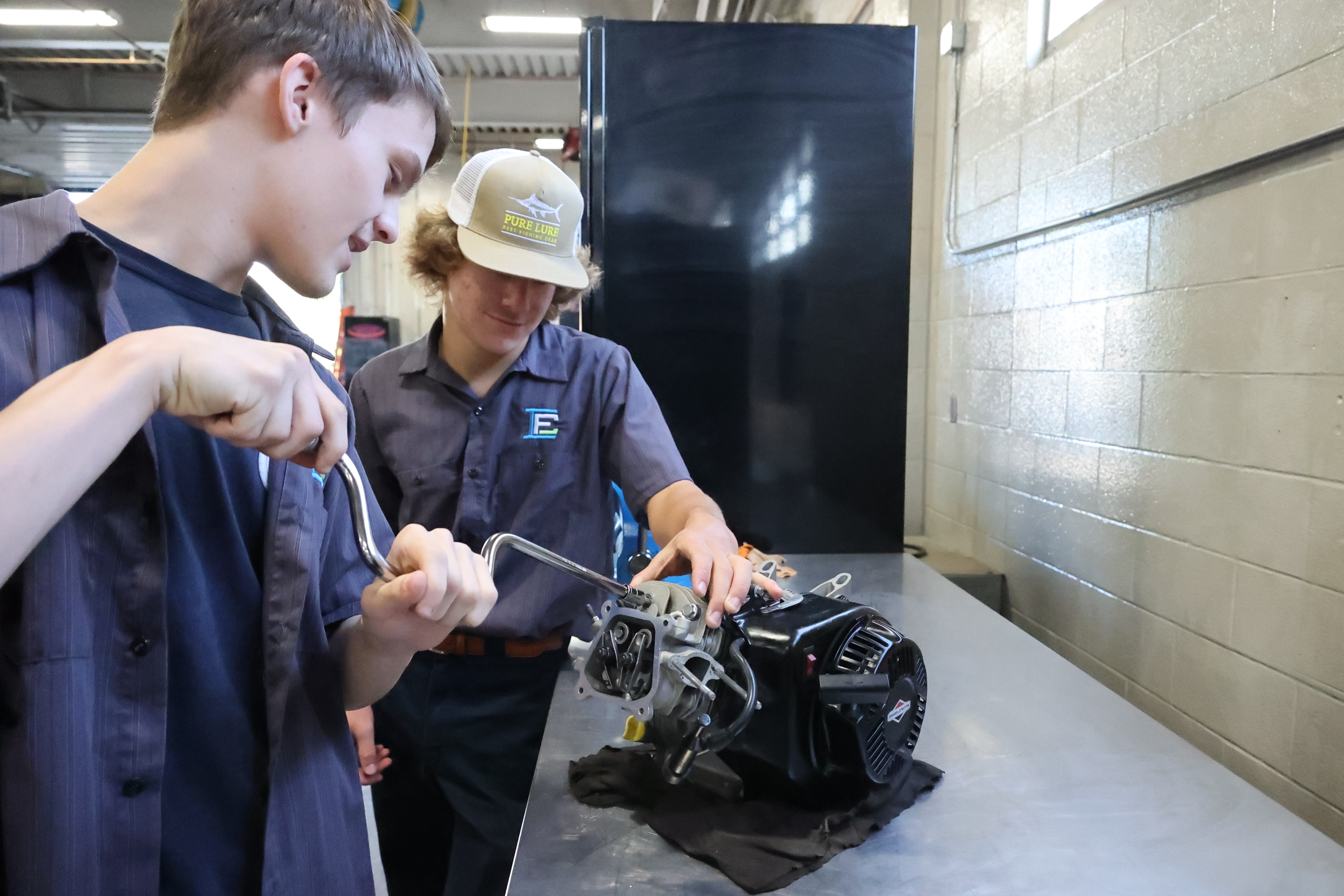 Two White, male students are working on an engine with a crank tool