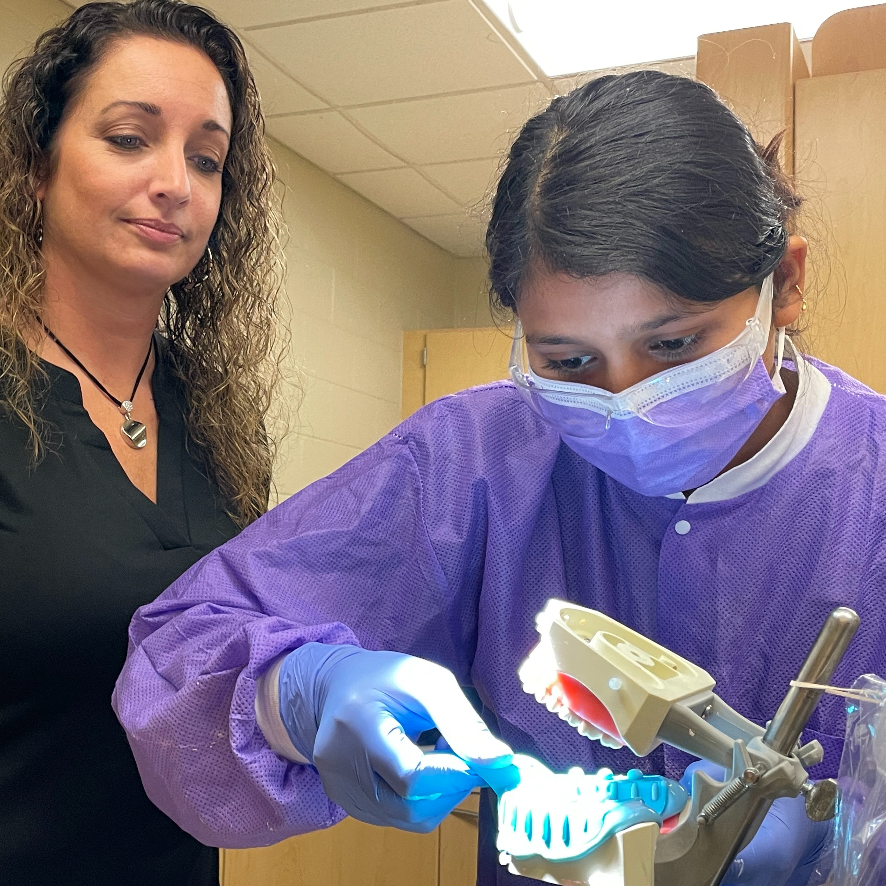 A woman of color is in purple scrubs and mask as she trains on cleaning a fake set of teeth.