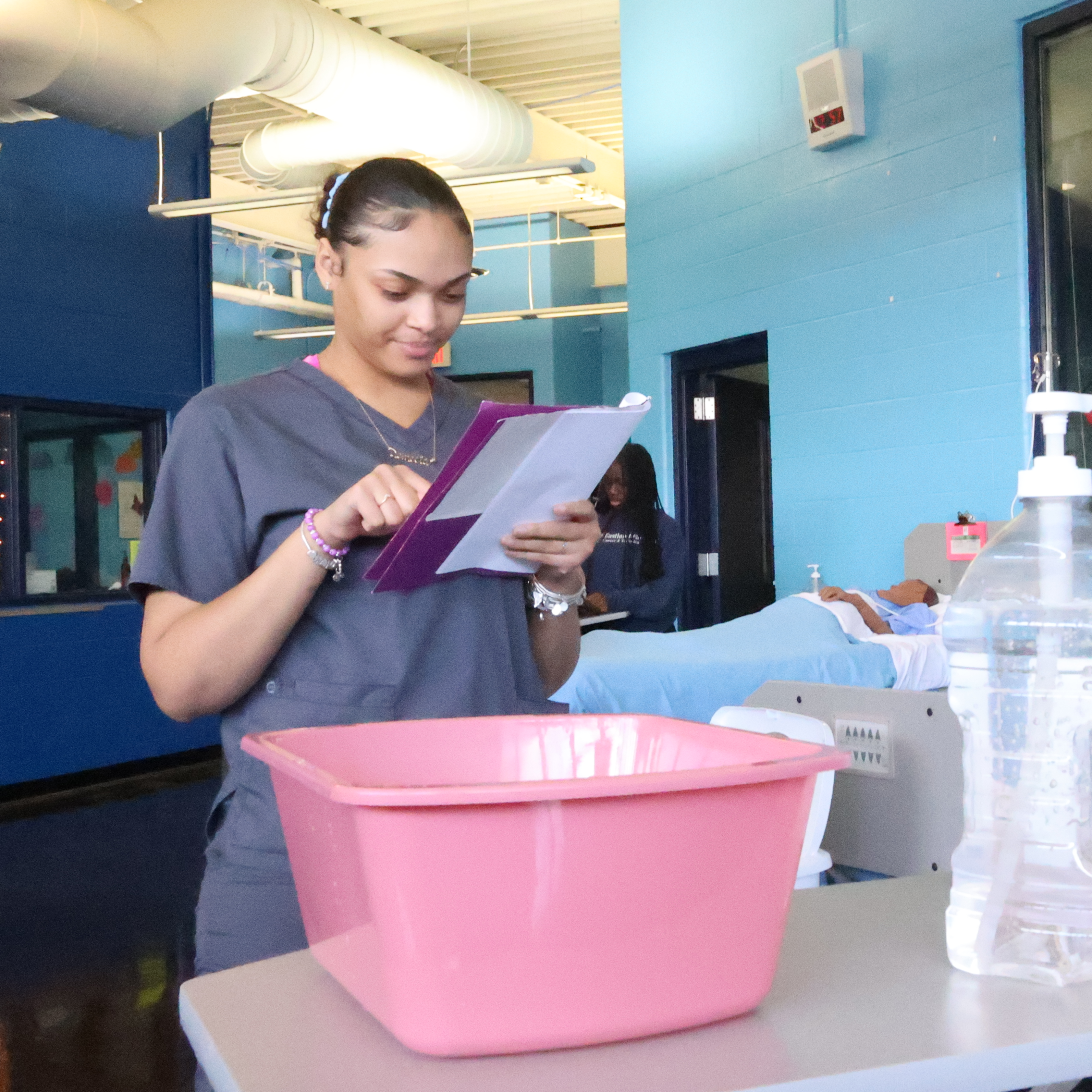 A Black, female student stands behind her cart of supplies that contains a bed pan and hand sanitizer. She is examining a checklist before giving patient care.