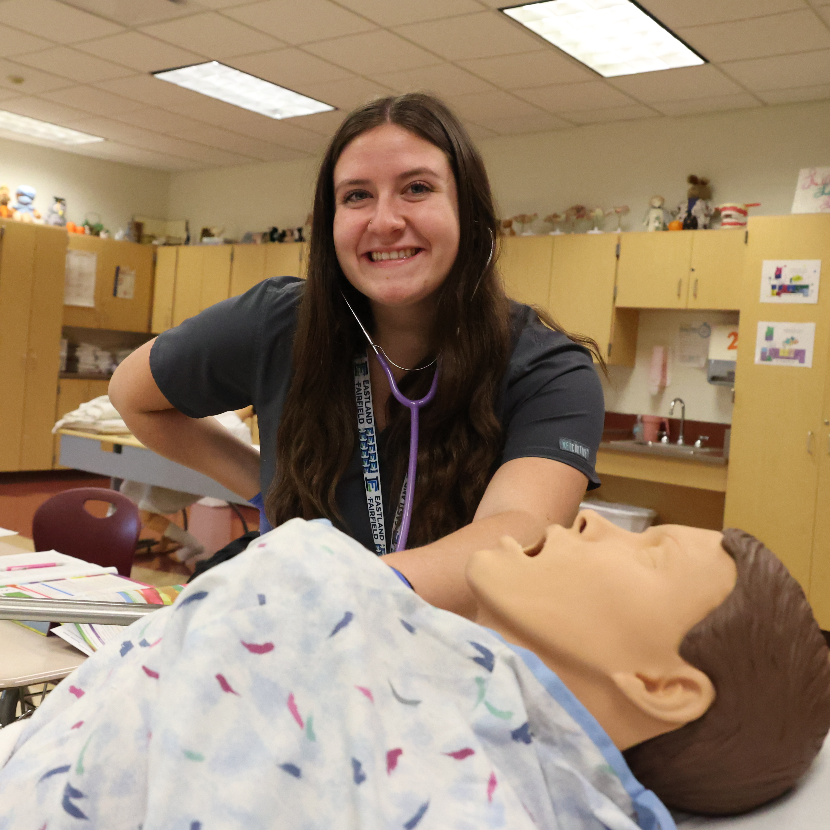 A White, female student is using a stethoscope to practice measuring the heartbeat of a patient on a mannequin.