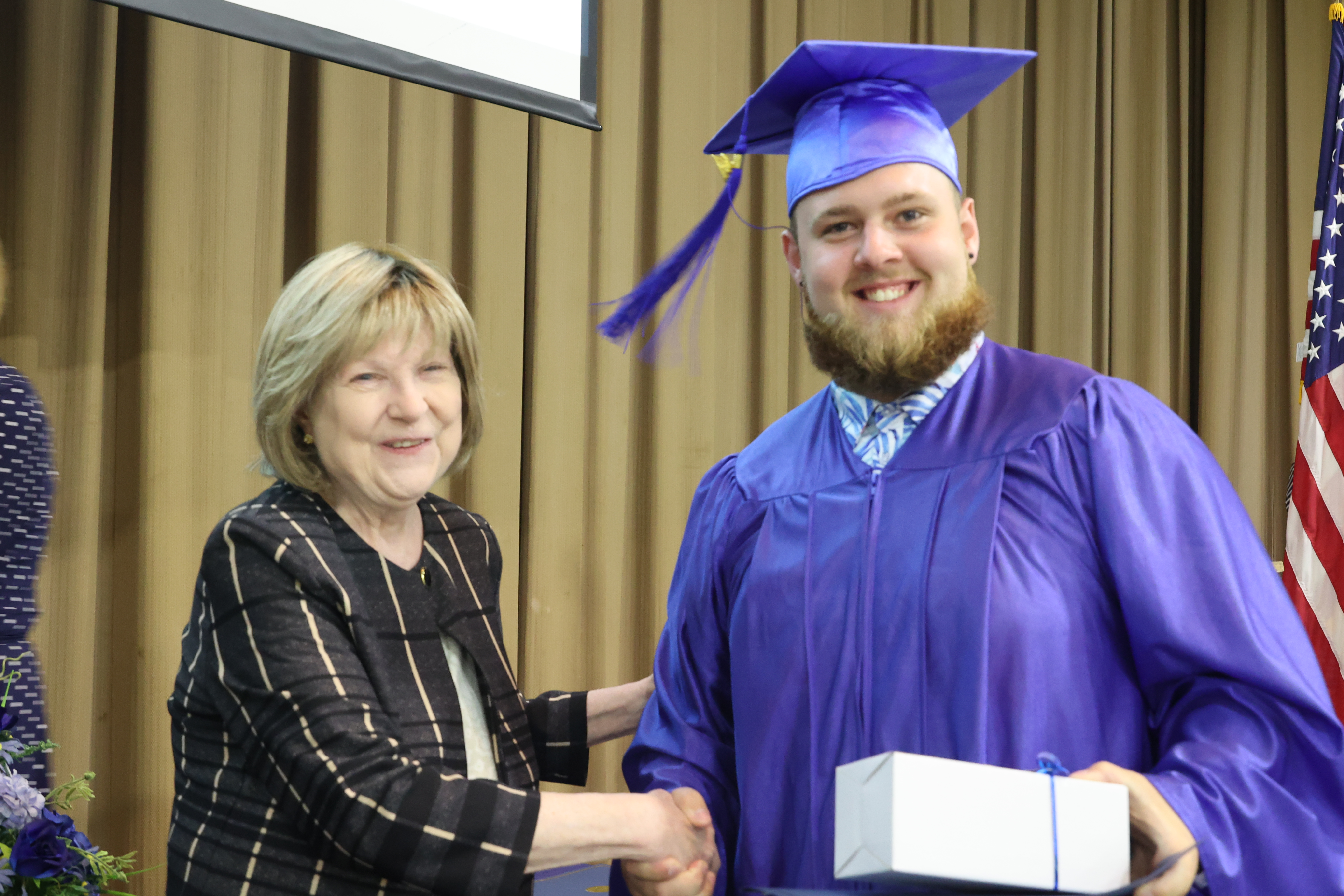 A White male student dressed in a blue cap and gown shakes the ASPIRE coordinators hand after receiving a gift.
