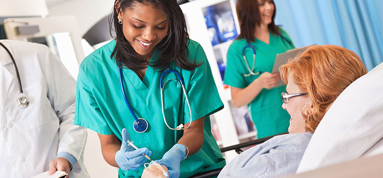 LPN stock image - a Black female nurse wearing green scrubs injects a patient with a needle