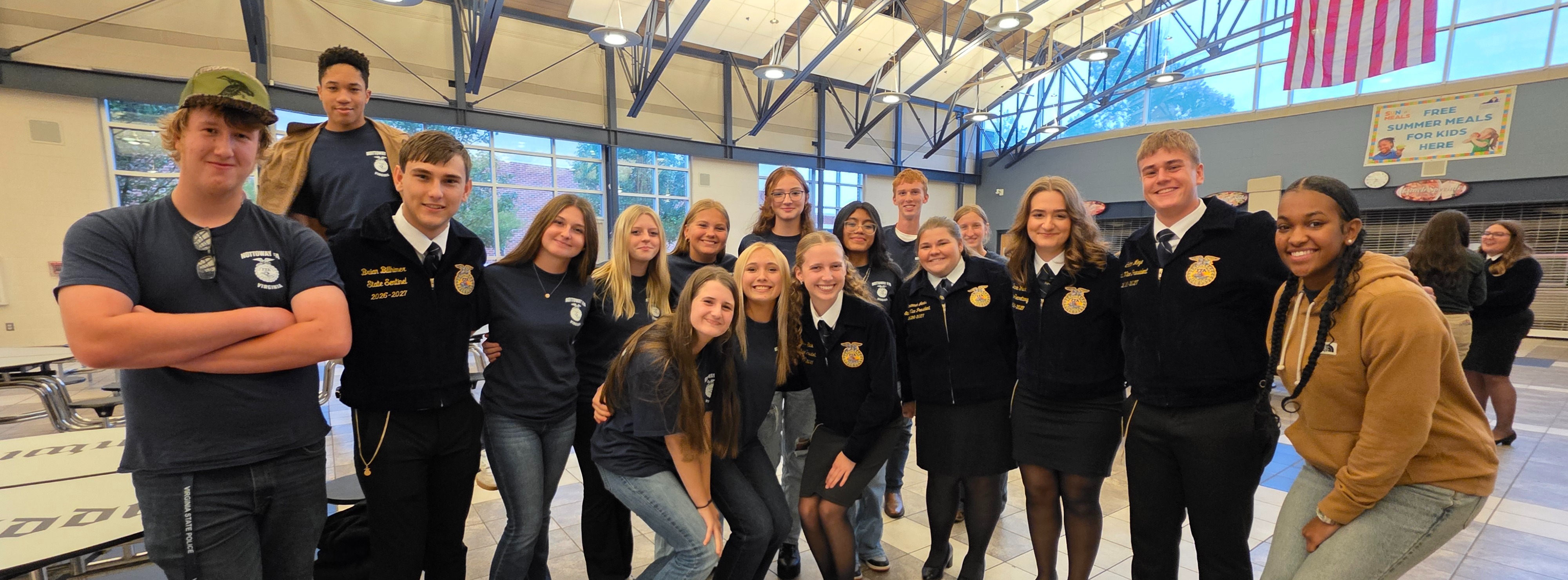 A group of students in a room with tiled flooring, wearing uniforms. An American flag is visible in the background.