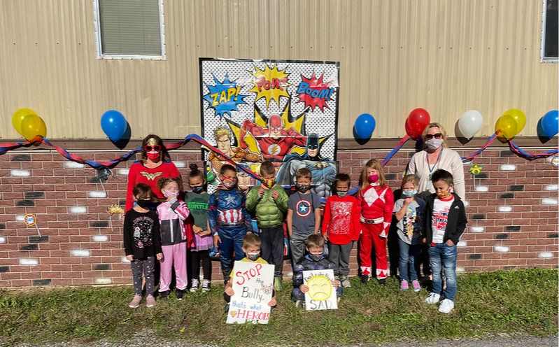 school counselors and students dressed up in superhero costumes holding "stop the bully that's what heroes do" sign in front of brick school wall decorated with a superhero poster and balloons
