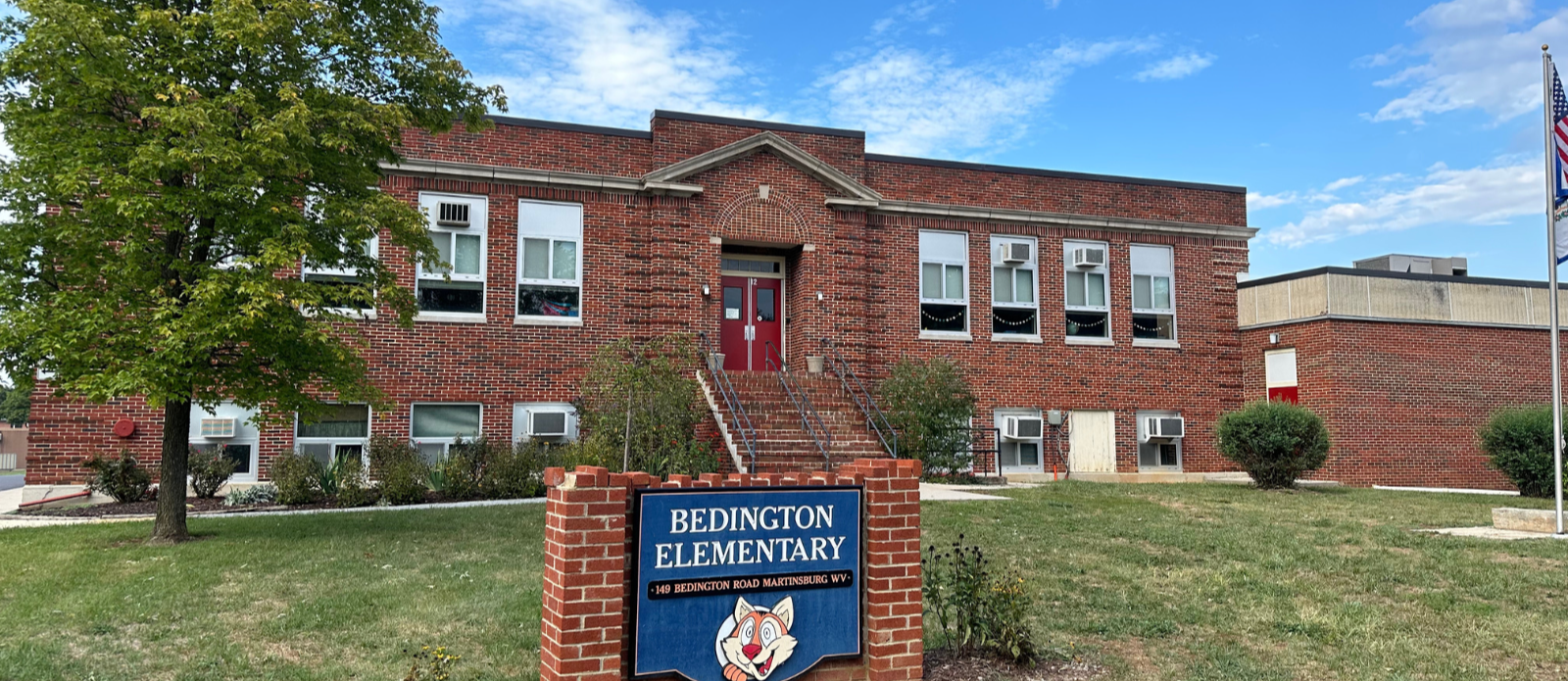 picture of the front of bedington elementary  - brick school building with red doors, grass lawn in front of it,  with blue bedington elementary sign on lawn