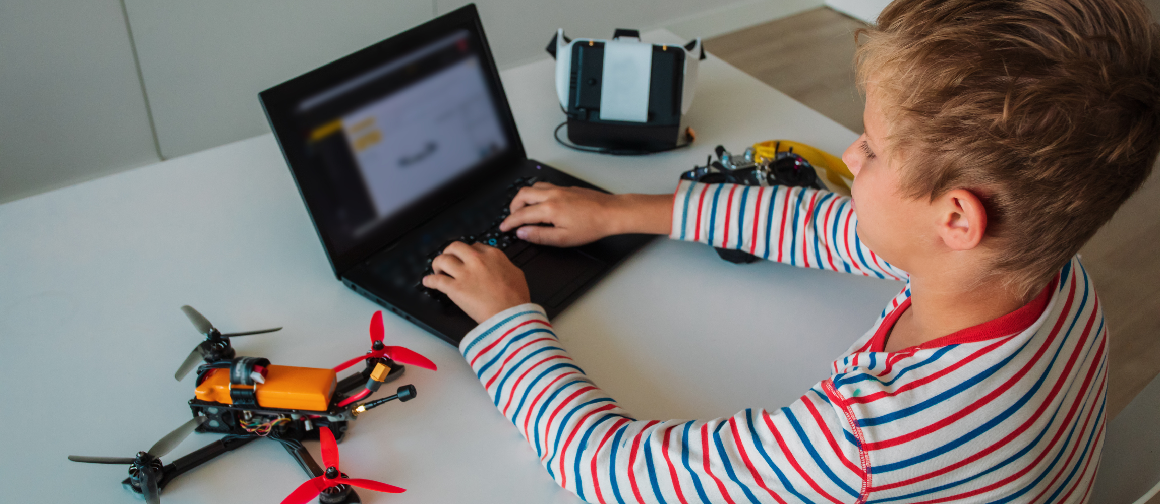 a boy types on a computer next to a drone and VR equipment