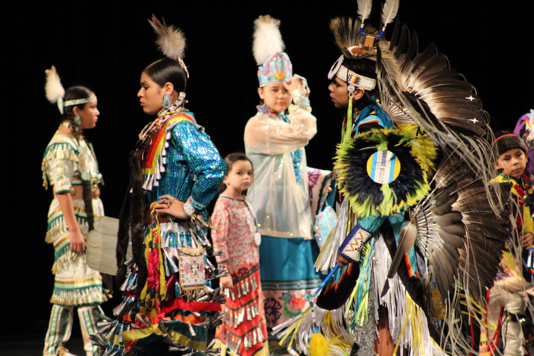 students wearing traditional tribal regalia dance on a stage