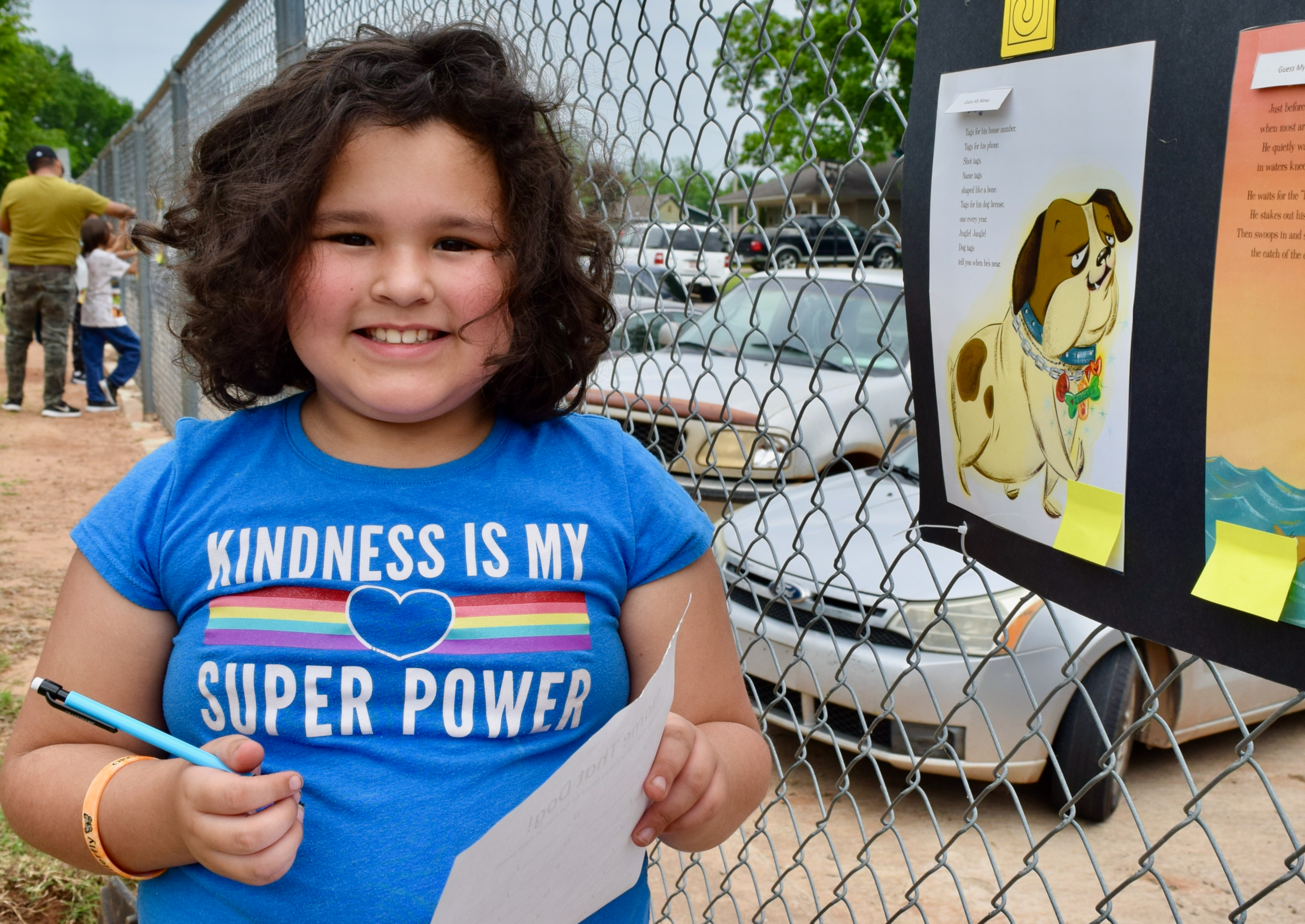 a girl wearing a shirt reading kindness is my super power stands next to a fence. she is holding a pencil and paper, and pages from a book are on the fence