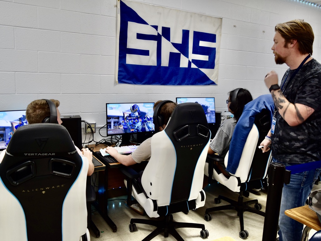 a man stands behind teenagers sitting in front of computers with a video game on the screen. an SHS banner is on the wall