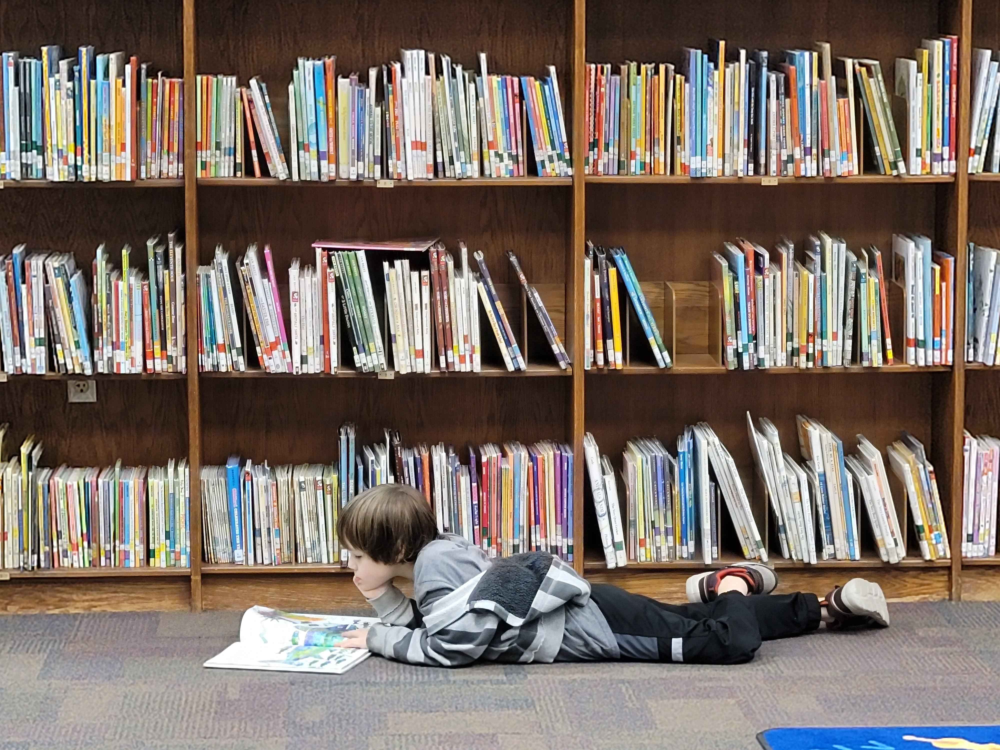 student laying on the carpet in front of a bookshelf reading