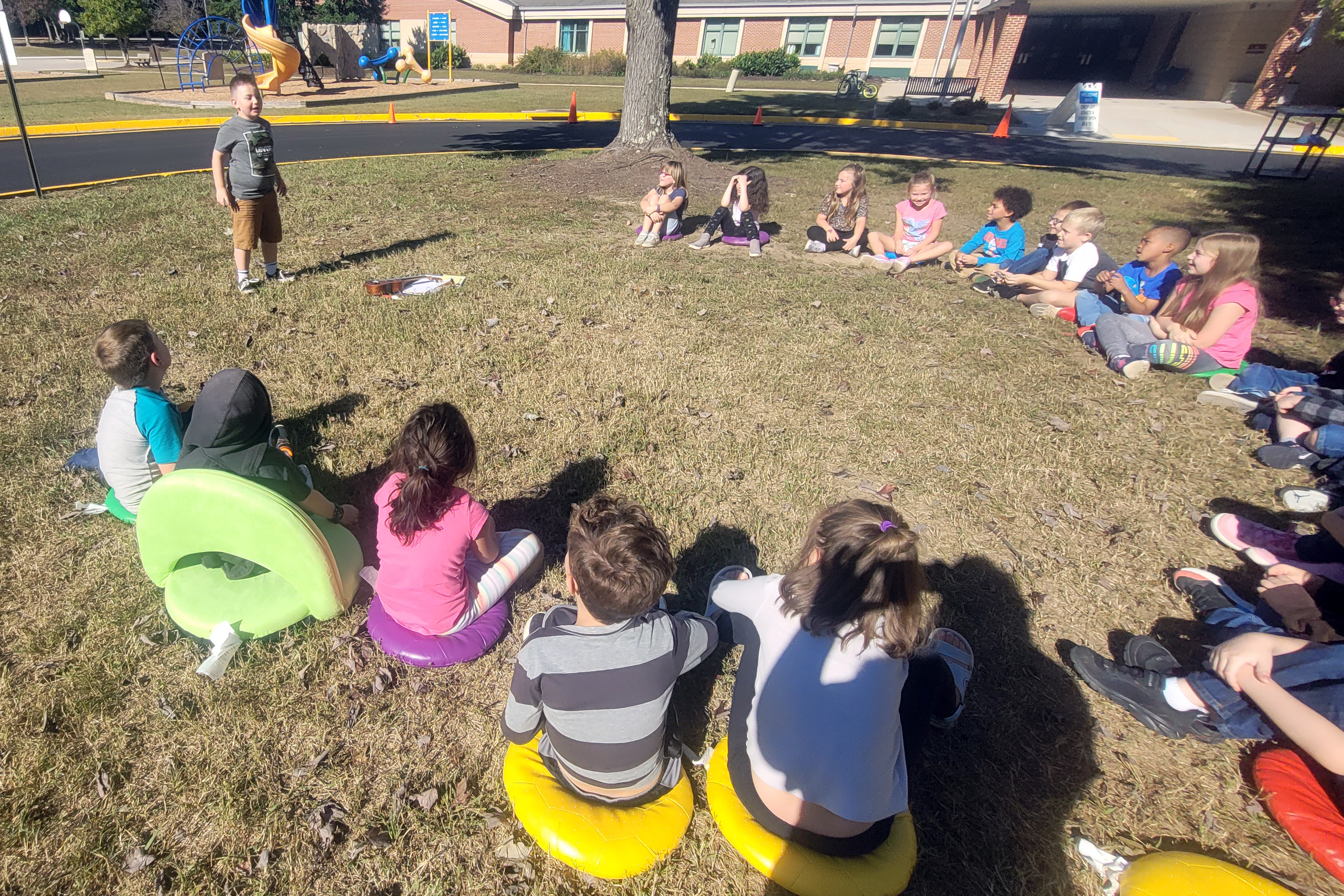 Large group of students in a circle on the ground. One standing up and talking.