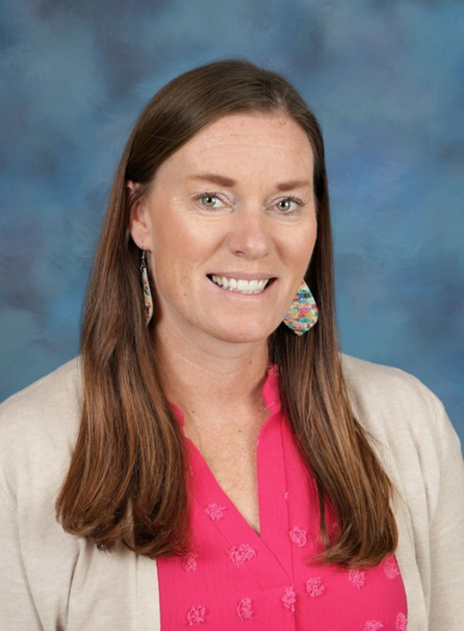 Woman with brown hair smiling at the camera, wearing pink blouse and a white cardigan. Background is blue.