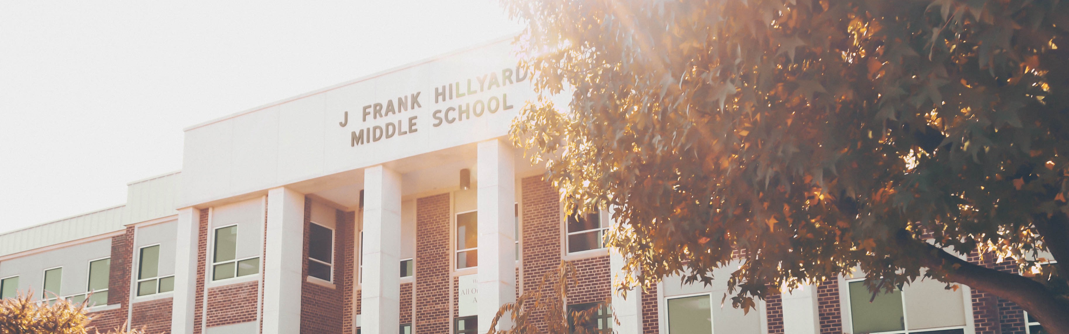 A white school building with columns, glass windows, and steps leading to the entrance. Green bushes and trees in front.