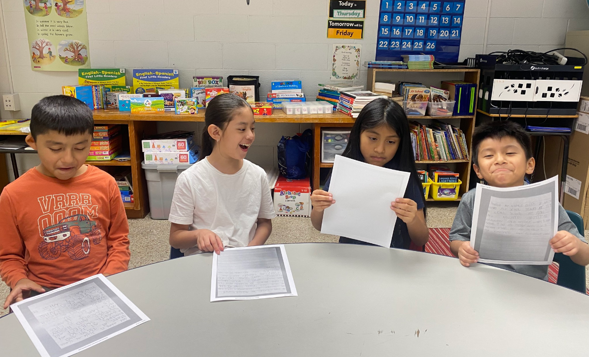 Four children hold papers, smiling in a classroom with bookshelves and a table in the background.