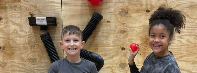 Two children in front of a wooden wall with equipment; one holds red objects and smiles, the other grins.