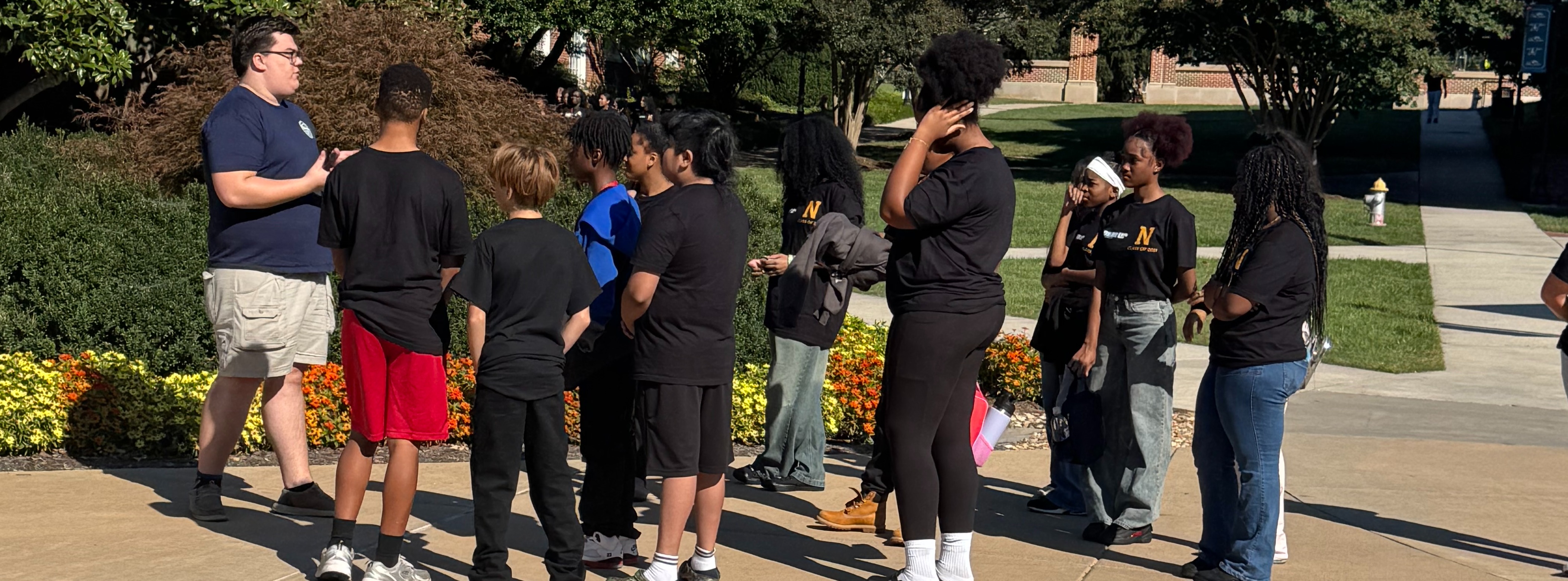 A group of people stands outside, conversing. They are on concrete near shrubs and trees.
