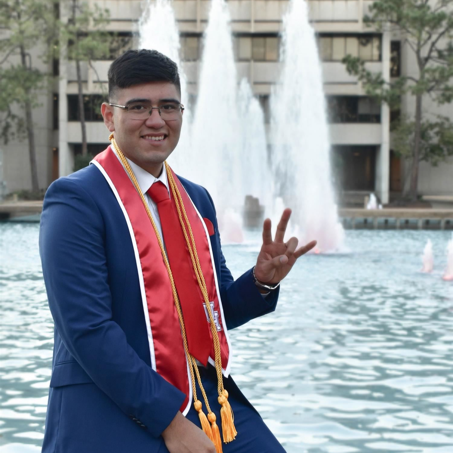 Miguel Lopez posing in front of an outdoor water fountain after graduation. Wearing business professional attire.