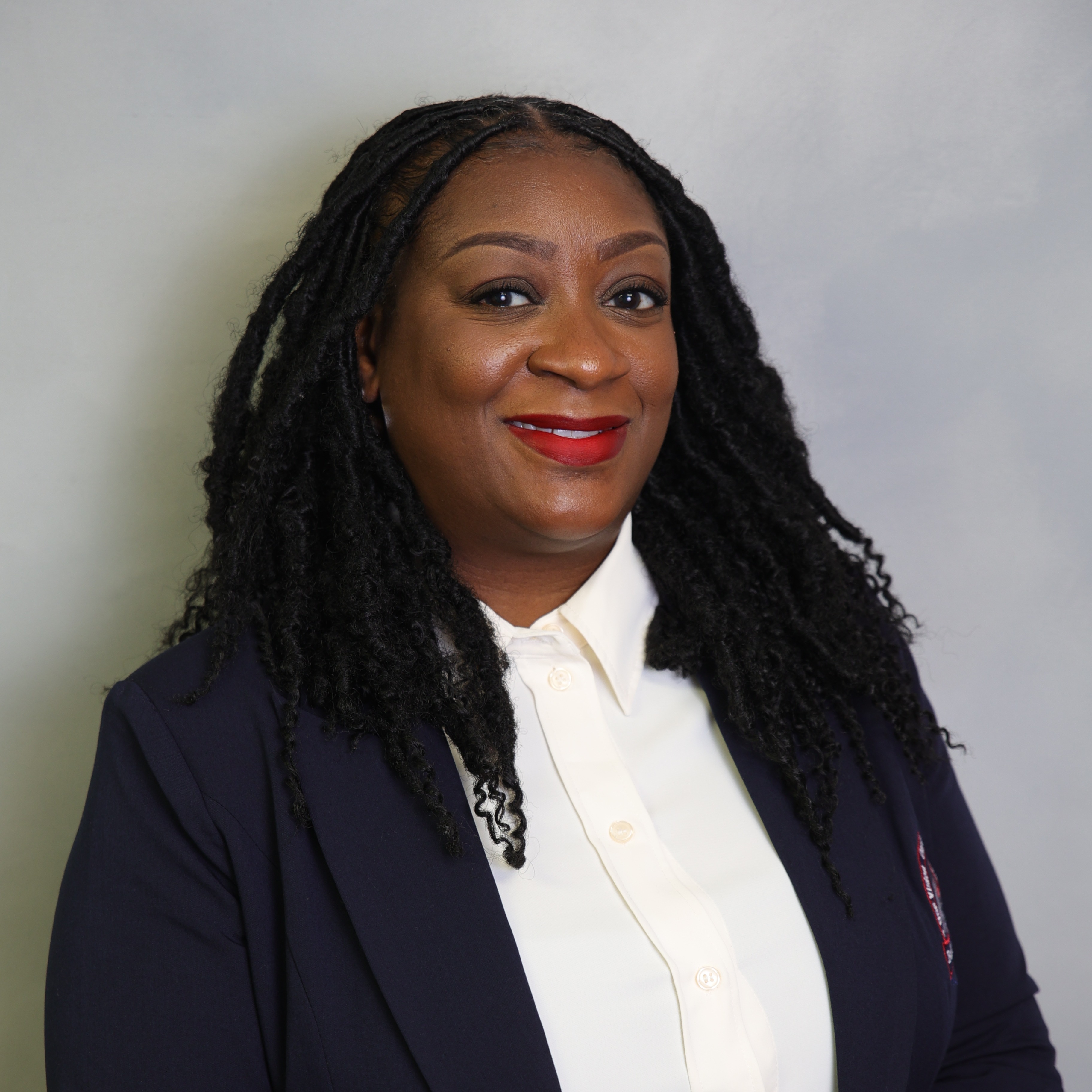 Professional portrait of Lorna Powell, Assistant Principal at Anthony Aguirre Junior High, smiling at the camera. She is wearing a navy blazer over a white collared blouse, with long black twists, against a light gray background.