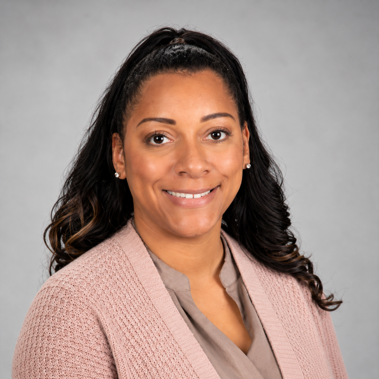 Professional headshot of Ms. Green, Social Studies Instructional Support Teacher (IST) at Anthony Aguirre Junior High. She is smiling and wearing a light pink cardigan over a taupe blouse. She has long, dark wavy hair styled half-up, small stud earrings, and is photographed against a light gray background.