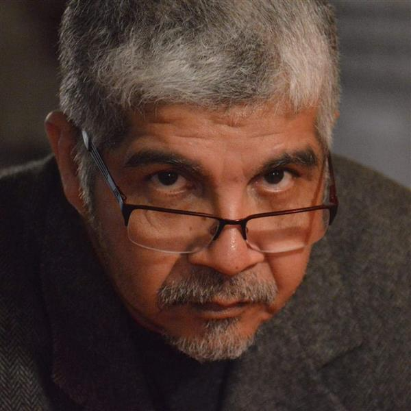 The image is a close-up portrait of Mr. Castro: Facial Features & Expression: He looks directly forward with a serious, intense gaze, peering through rectangular dark-framed glasses that rest slightly low on the bridge of his nose. He has dark, defined eyebrows and dark eyes. Hair & Facial Hair: He has short, salt-and-pepper gray hair and a neatly trimmed graying mustache and goatee. Attire: He is dressed in a dark inner shirt beneath a dark gray, textured tweed or woolen jacket. Composition & Lighting: The photo is tightly focused on his face and shoulders against a dark, softly blurred background under warm lighting.