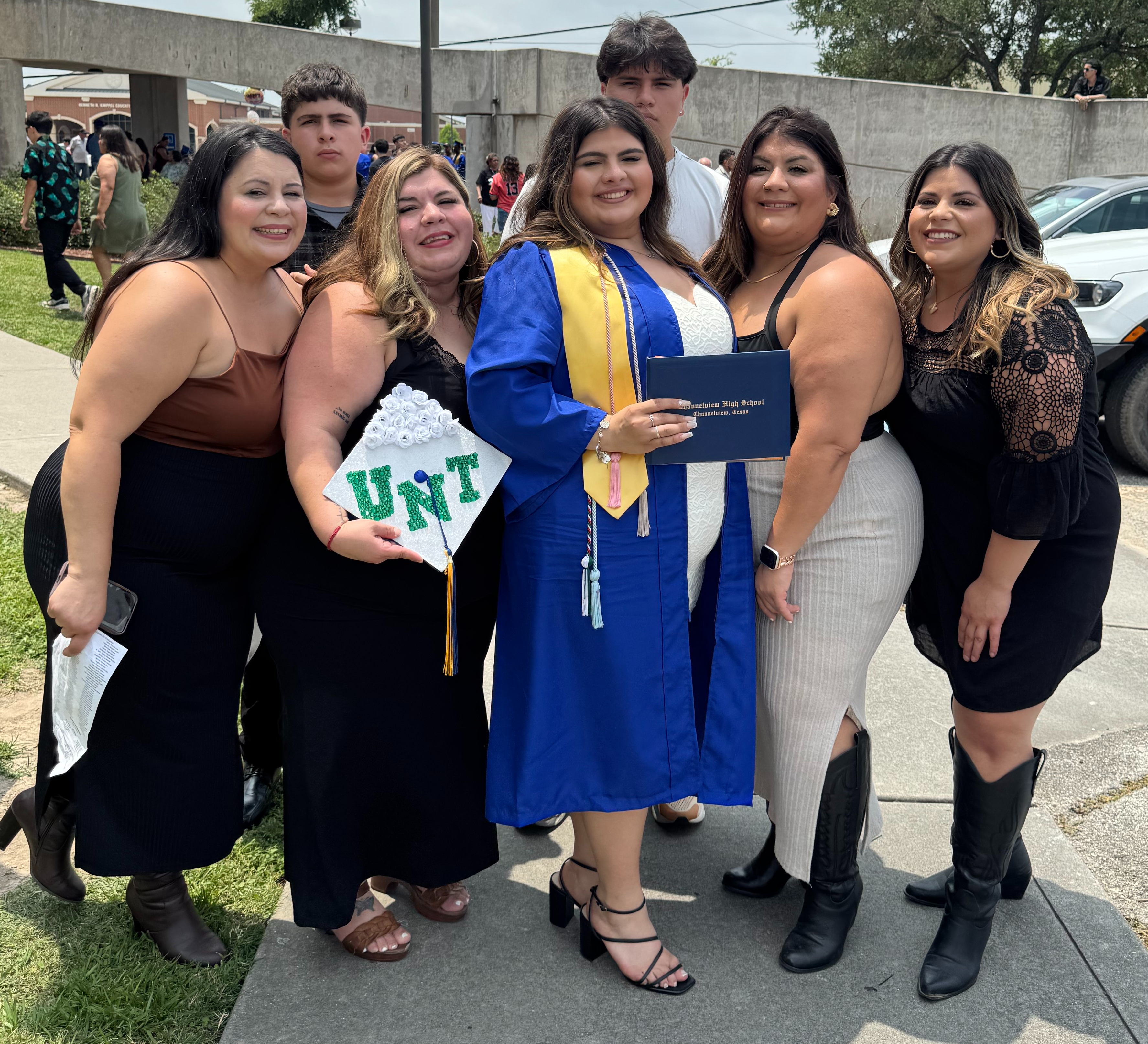 “A graduate in a blue cap and gown with a gold stole holds her diploma while posing outdoors with family.”