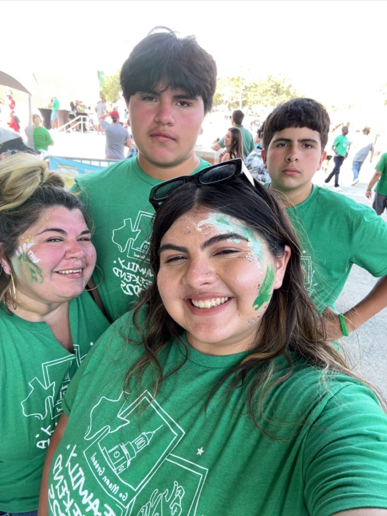 “Four people wearing matching green shirts pose for an outdoor selfie, with green and white face paint on the two people in front at a football game.”Family Weekend at UNT!!!