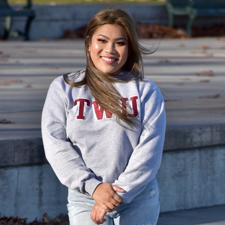 The photograph is a portrait of Elizabeth Keomanikhoth, a young woman standing outdoors on a set of wide, gray concrete steps. She is positioned near the center of the image, facing the camera with a warm, confident smile. Her posture is relaxed and natural, giving the photograph a friendly, approachable feeling. Her face and expression: Elizabeth has a broad, genuine smile that shows her upper teeth. Her cheeks are slightly raised, and her eyes appear bright and gently narrowed with her smile. She has neatly shaped eyebrows, dark eye makeup that subtly accentuates her eyes, and a warm, natural-looking complexion. Her makeup is polished but not overly dramatic, with a soft glow on her cheeks and a rosy, glossy finish on her lips. Sunlight illuminates her face, especially her cheeks and forehead, creating a soft highlight. Her hair: Her hair is long, straight, and predominantly light-to-medium brown, with lighter caramel and golden-brown highlights that catch the sunlight. It has a subtle side part near the top of her head. Most of her hair falls over her shoulders and down the front of her sweatshirt, with a large section draped diagonally across her chest. A few loose strands are swept toward the right side of the photograph, suggesting a gentle breeze. Her hair reaches approximately to her mid-chest in the front. Her clothing: Elizabeth is wearing a light gray, long-sleeved crewneck sweatshirt with a relaxed fit. Across the chest is the word “TWU” in large, bold, red collegiate-style letters outlined in white. The letters stand out prominently against the pale gray fabric. The sweatshirt has ribbed cuffs and a ribbed waistband, and the sleeves gather slightly around her wrists. She pairs it with light-wash blue jeans. The jeans have a casual fit and are visible from the waist down to the upper thighs, where the photograph ends. Her pose and body language: Elizabeth stands upright with her shoulders relaxed and her body angled just slightly rather than completely square to the camera. Both arms are bent gently in front of her body. Her hands are clasped loosely together at about hip level, with one hand resting over the other. Her fingers are relaxed rather than tightly interlocked. This simple pose gives the portrait a comfortable, composed appearance. She looks directly toward the camera, making the image feel personal and engaging. The background and surroundings: Behind Elizabeth are several broad, horizontal concrete steps in shades of gray. The steps have slightly weathered surfaces, with a scattering of small brown leaves along the edges, suggesting an autumn setting. At the top of the photograph, the background becomes softer and more out of focus. There are dark blue-gray structural elements and a few indistinct objects, possibly parts of an outdoor campus seating area or nearby architecture. The background is understated and does not distract from Elizabeth. The lighting and overall atmosphere: Natural sunlight falls across the scene, brightening Elizabeth's face, hair, and sweatshirt. The light creates warm golden highlights in her hair and gentle shadows beneath her chin and along the folds of her clothing. The image is sharpest around her face and upper body, while the background is softly blurred. The overall color palette combines cool grays and muted blues with the warm brown tones of her hair, the red lettering on her sweatshirt, and her sunlit complexion.