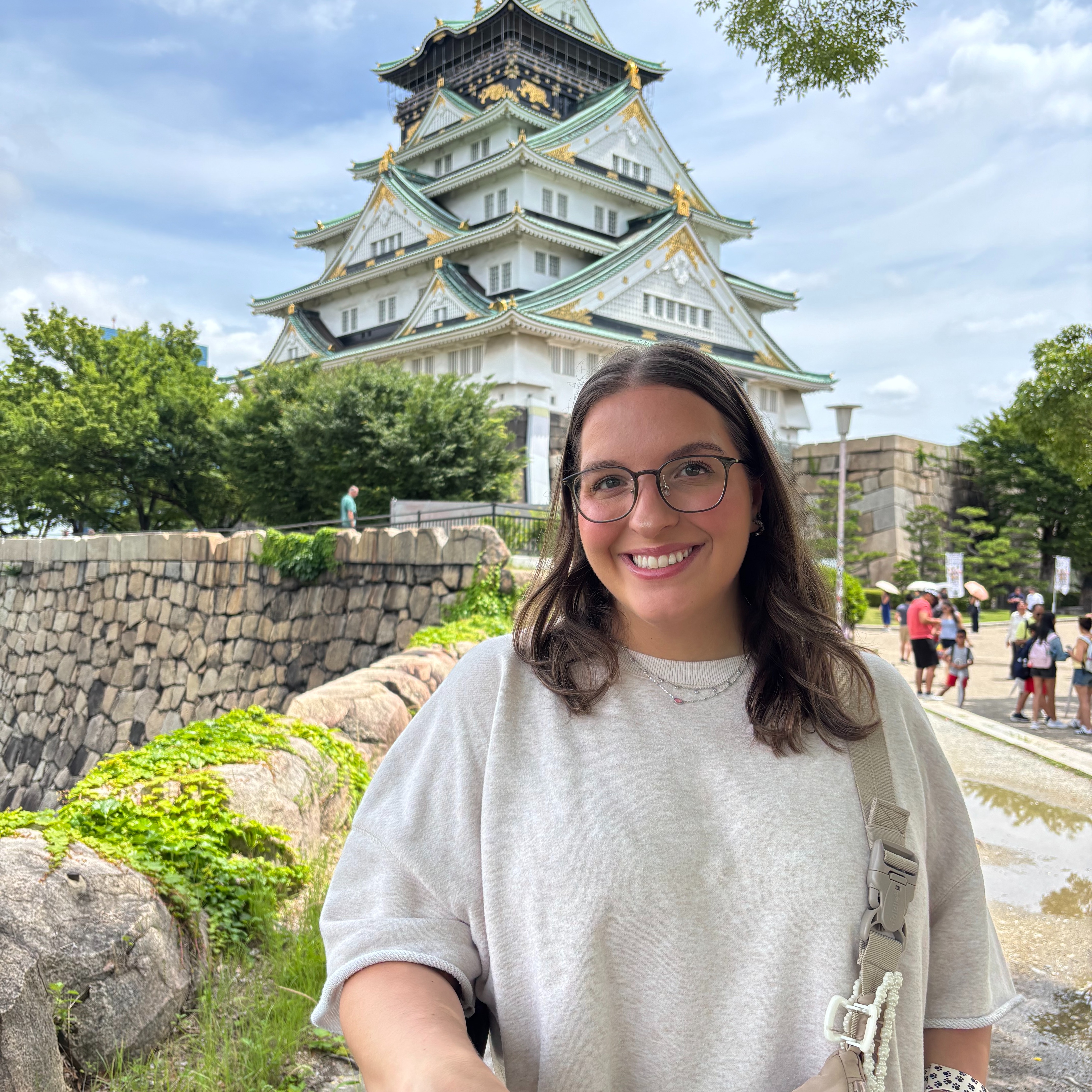 The photograph shows a smiling woman standing outdoors in front of a traditional Japanese castle on a bright, pleasant day. In the foreground, a woman stands slightly right of center, facing the camera and smiling warmly with her teeth visible. She has shoulder-length, dark brown hair parted slightly off-center, with soft waves at the ends. She wears round, dark-framed eyeglasses and has a light complexion. She is dressed casually in a loose-fitting, light gray crewneck sweatshirt. A small, delicate necklace is visible around her neck. A wide, beige crossbody bag strap runs diagonally over her left shoulder, with a light-colored plastic buckle near her chest. Her arms are relaxed, and one forearm is visible near the bottom of the photograph. Behind her, filling much of the upper half of the image, stands a large, traditional Japanese castle. The castle has multiple stacked levels with white exterior walls, dark wooden accents, and gracefully curved, dark gray tiled roofs. The edges of the roofs feature decorative gold and muted green details. Each level becomes progressively smaller toward the top, creating a tall, tiered silhouette. The highest section has an ornate, dark roof with golden accents and a small decorative finial at its peak. The castle's white walls contrast with its dark rooflines, giving the building a striking, elegant appearance. At the base of the castle is a substantial stone wall made of irregularly shaped, gray and tan stones. The wall extends from the left side of the photograph toward the center, behind the woman. In the lower-left foreground, several large, rounded rocks sit beside the wall, surrounded by bright green ground cover and low plants. A few leafy trees frame the scene on both sides, with dense green foliage particularly visible along the left edge. On the right side of the photograph, a paved pedestrian walkway extends into the background. Several distant visitors are walking along it, including people wearing colorful clothing, such as red, blue, and light-colored tops. These figures are much smaller than the woman in the foreground, emphasizing the depth of the scene and the scale of the castle. Above the castle, the sky is pale blue with soft, wispy white clouds. Natural daylight illuminates the scene evenly, highlighting the woman's face, the castle's bright white walls, and the surrounding greenery without creating harsh shadows. Overall impression: The photograph captures a cheerful travel moment. The woman is the clear focal point in the foreground, while the magnificent Japanese castle, historic stonework, lush greenery, and visitors in the background establish a scenic cultural landmark as the setting. The composition feels relaxed and inviting, with the woman positioned prominently against the impressive architecture.