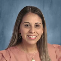 The picture shows Brenda Garcia smiling warmly at the camera for a professional portrait. She has long, straight brown hair with lighter highlights, parted in the middle. She has brown eyes, neatly shaped eyebrows, and a friendly smile showing her teeth. She is wearing a dusty rose-colored blouse, small dangling earrings, and a delicate gold necklace with a pendant. The background is a soft blue-gray studio backdrop.  Overall, the portrait presents Brenda as friendly, approachable, and professional, making it suitable for a school staff directory, teacher profile, or professional biography.