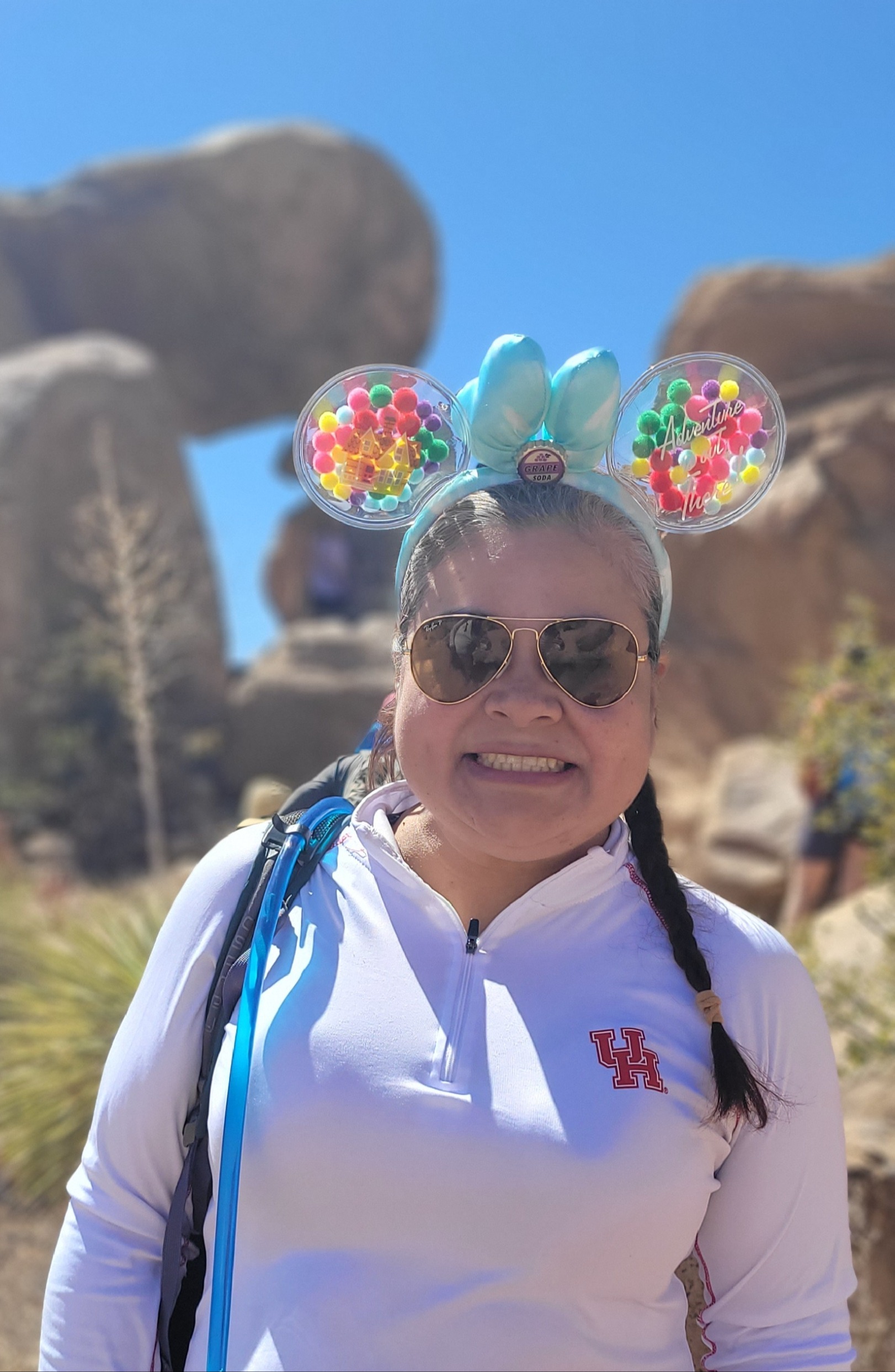 Mrs. M. Lopez is the first grade teacher featured in this photo. She is the main focus of the photograph. She is positioned in the center of the image, visible from approximately the waist up. She has medium-toned skin and dark hair with some gray visible near the front and along her hairline. Her hair is pulled back, with a long, dark braid draped over her left shoulder (on the right side from the viewer’s perspective). She is wearing large, dark-tinted aviator sunglasses with thin, gold-colored frames. Her face has a broad, cheerful smile, with her teeth showing, giving her a friendly and enthusiastic appearance.  On her head, she wears a playful pair of oversized, mouse-ear-style headwear. The two large, round, transparent ears are positioned on either side of her head, with bright, multicolored decorations inside them, including small spheres in shades of pink, red, yellow, green, blue, and purple. Two tall, light-blue shapes resembling mouse ears or bows rise between the round decorations. The right circular ear, from the viewer’s perspective, features decorative lettering that reads “Adventure Awaits,” adding a fun, travel-themed touch to her outfit.  She is dressed in a light lavender or pale purple long-sleeved athletic pullover. The shirt has a short zipper at the front of the collar and a small red-and-white University of Houston “UH” logo on the left side of her chest, which appears on the right side from the viewer’s perspective. A black backpack strap with bright blue trim runs over her right shoulder, visible on the left side of the photograph. The strap suggests that she may be carrying a backpack while exploring the area.  The background features a striking rocky landscape. Behind the woman, enormous rounded rock formations rise on both sides of the image. The rocks are in shades of warm brown, rusty red, tan, and muted gray, with uneven surfaces, weathered contours, and deep crevices. On the left side of the photograph, a massive rock formation has a broad, rounded upper section that extends horizontally, creating a shape somewhat reminiscent of an arch or an overhang. On the right, another large, reddish-brown rock formation curves upward, framing the woman and helping create a sense of depth.  The sky fills the upper portion of the background and is a brilliant, cloudless blue. The strong sunlight illuminates the woman’s face and clothing, producing bright highlights on her shirt and sunglasses, along with distinct shadows beneath her chin and around the folds of her clothing.  Near the bottom and sides of the photograph, small patches of desert-like vegetation are visible. These include clusters of long, narrow, pale-green and yellow-green leaves growing close to the ground.