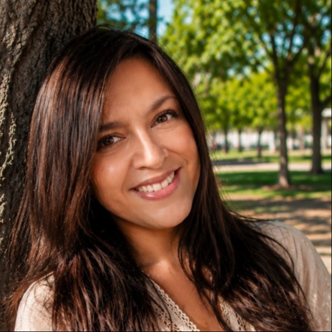 A close-up portrait of teacher Mrs. Merari Gonzalez. She is smiling warmly at the camera, leaning against a tree trunk in a bright, sunlit park. She has long, dark brown hair and is wearing a cream-colored blouse with lace detailing along the neckline. The background shows blurred green trees and a park walkway.