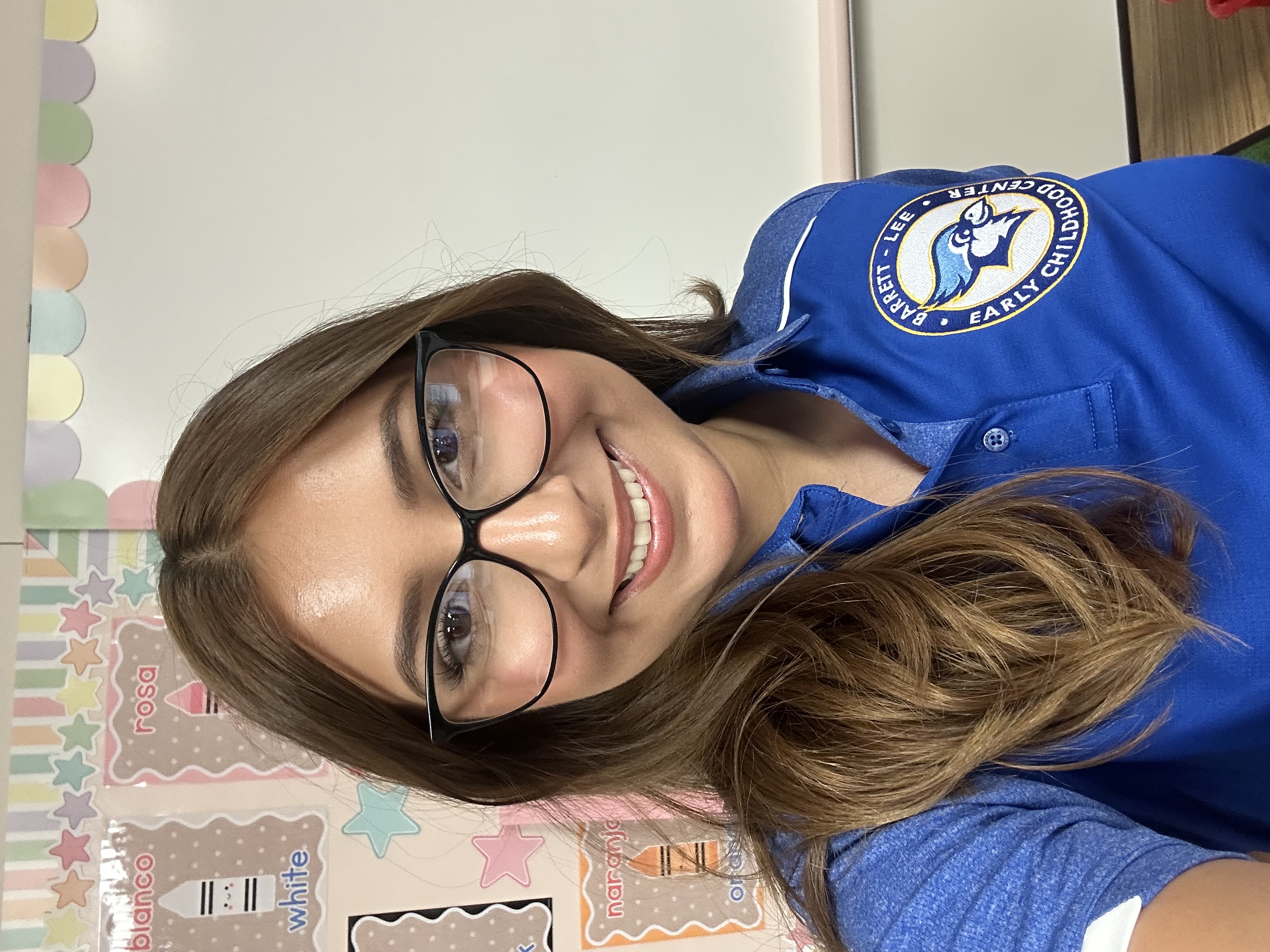 Smiling young woman with long brown hair and black glasses poses for a picture in a classroom. She is wearing a blue polo shirt with a Berrett Lee Early Childhood Center logo.