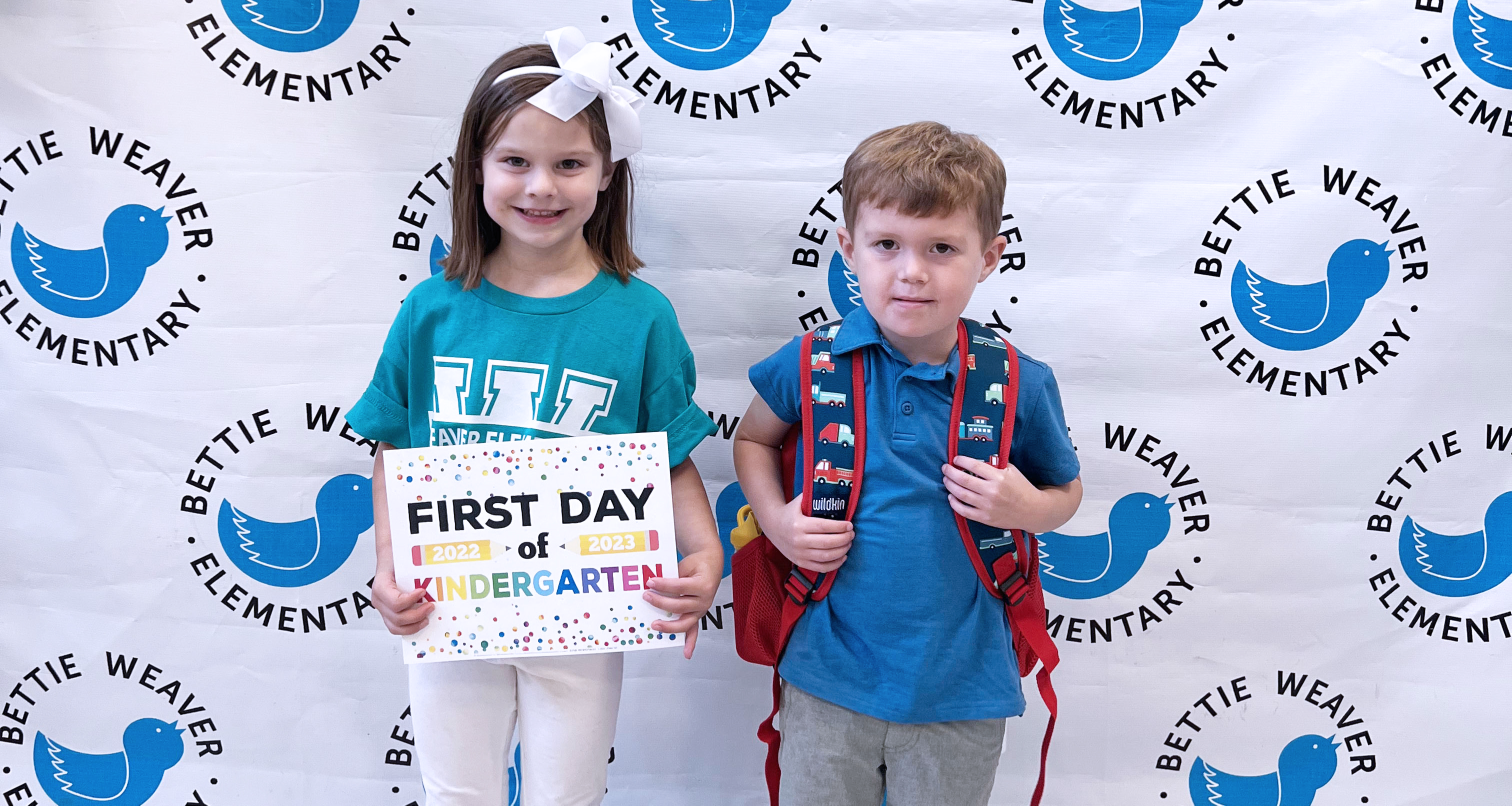 Two students in front of step and repeat wall holding first day sign.
