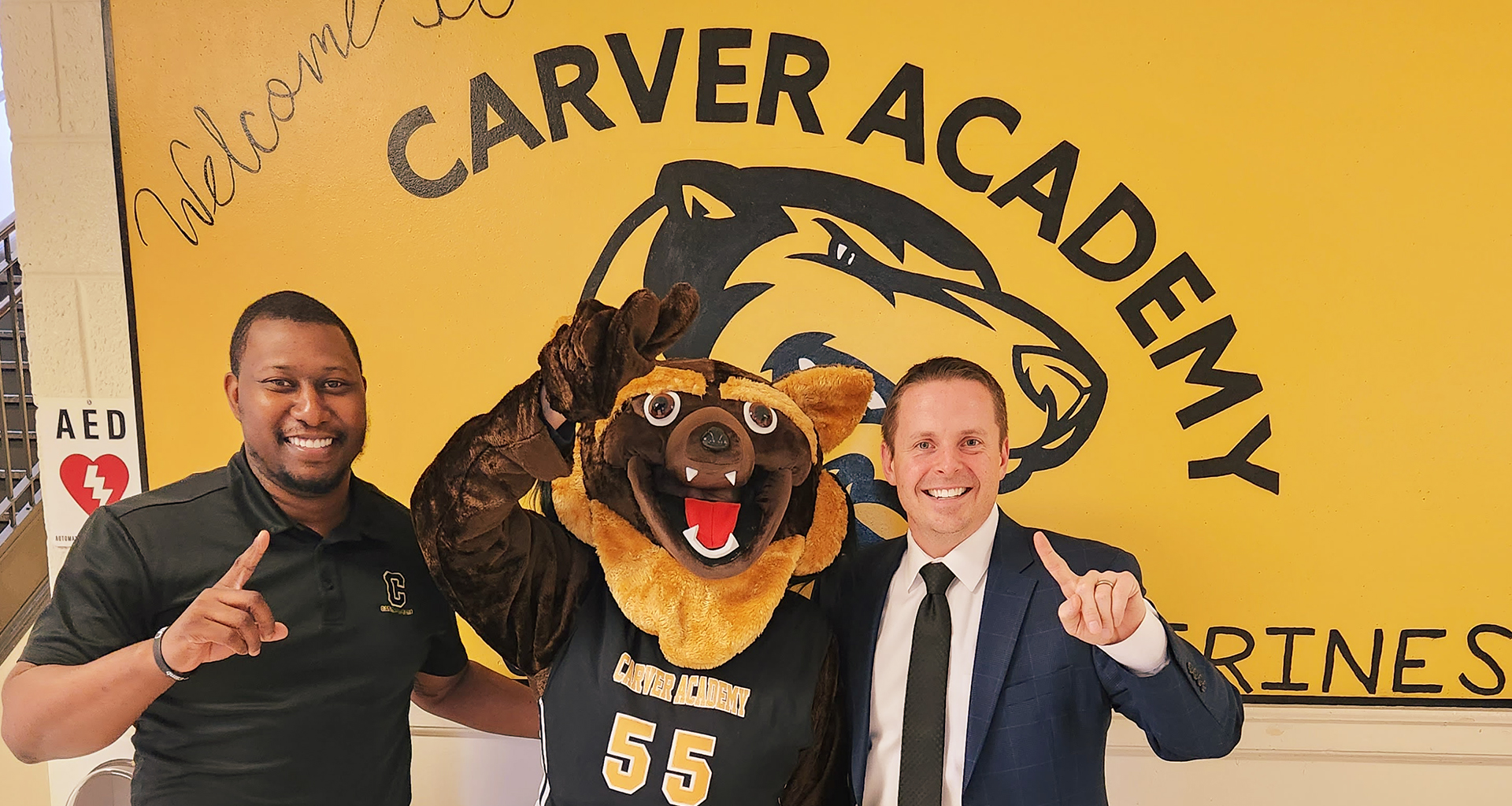 Principal and AP pose with mascot in front of welcome sign