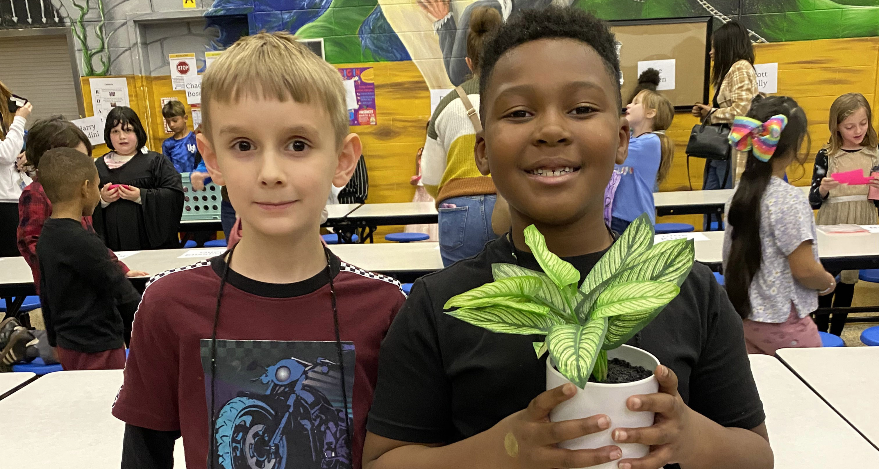 Two students in the cafeteria while one holds a small plant